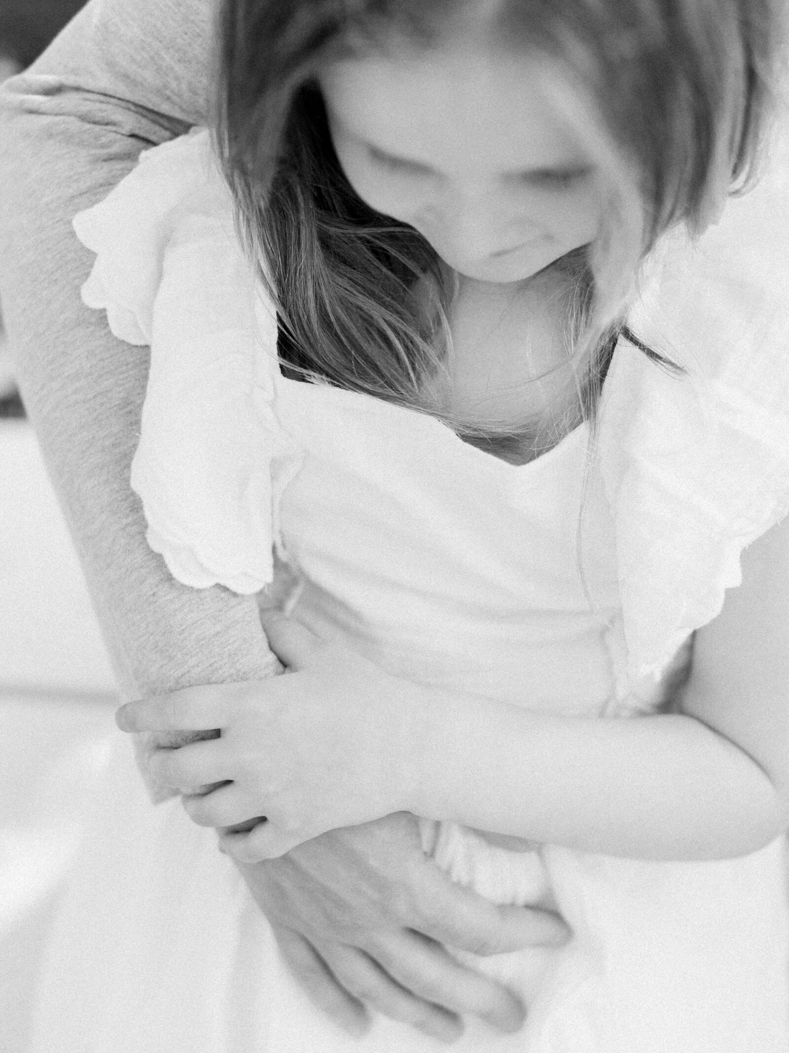 Closeup black and white photo of little girl in a white linen dress, holding onto her dad's arms by NH newborn photographer Fieldstone Studio.