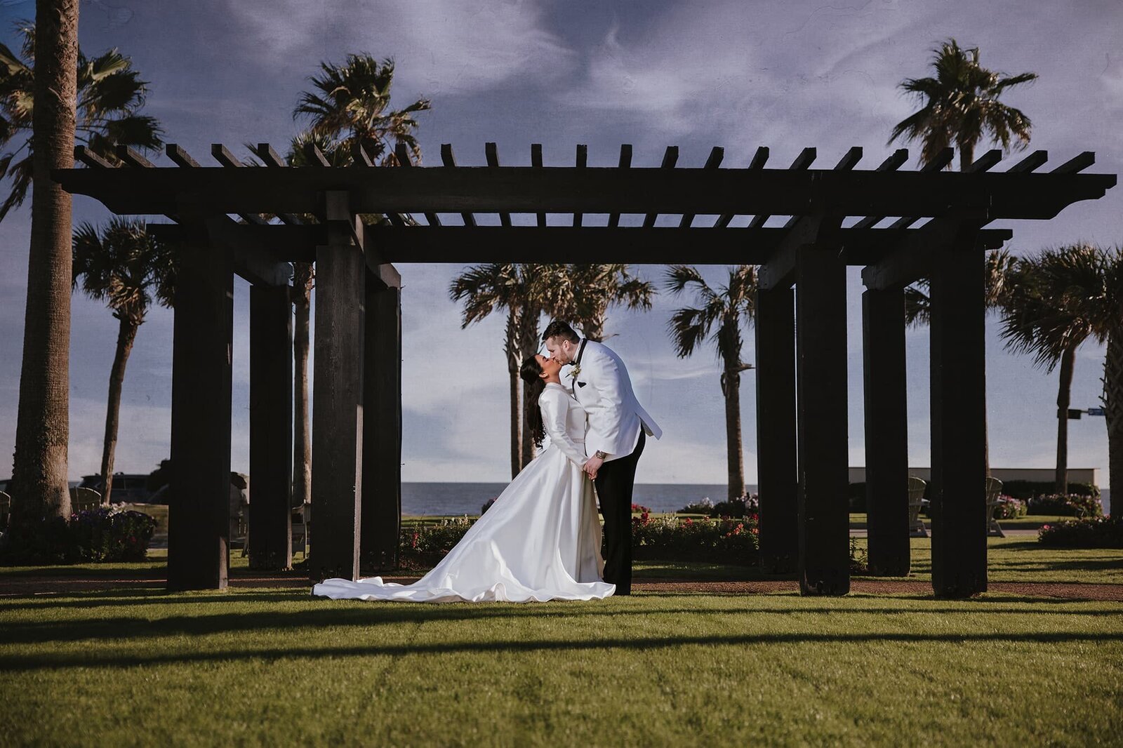 Romantic beachfront wedding portrait at Grand Galvez in Galveston, TX, featuring a bride and groom sharing a kiss beneath a black pergola, framed by palm trees and ocean views — captured by luxury wedding photographer Logrono & Co. Weddings.