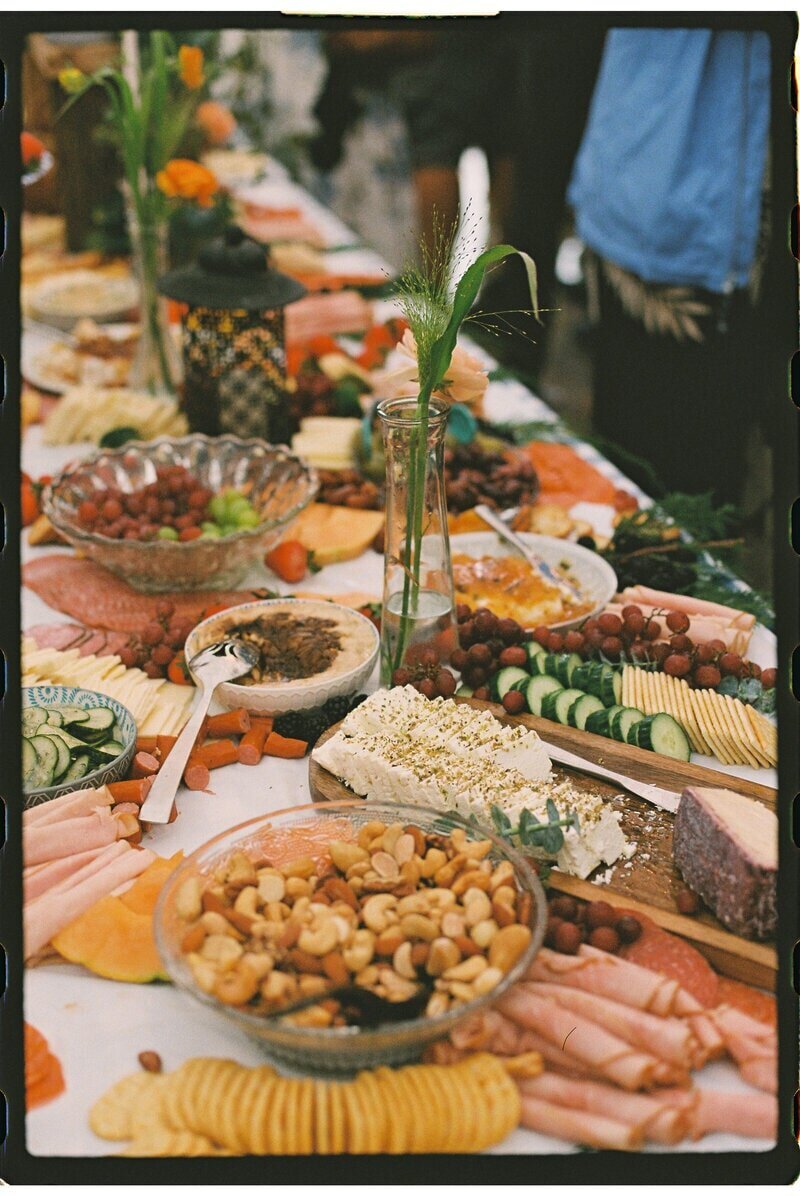 A table filled with an assorted spread, including cheese, meats, nuts, crackers, sliced cucumbers, and grapes. There are decorative plants and flowers placed among the food, creating an inviting and colorful display.