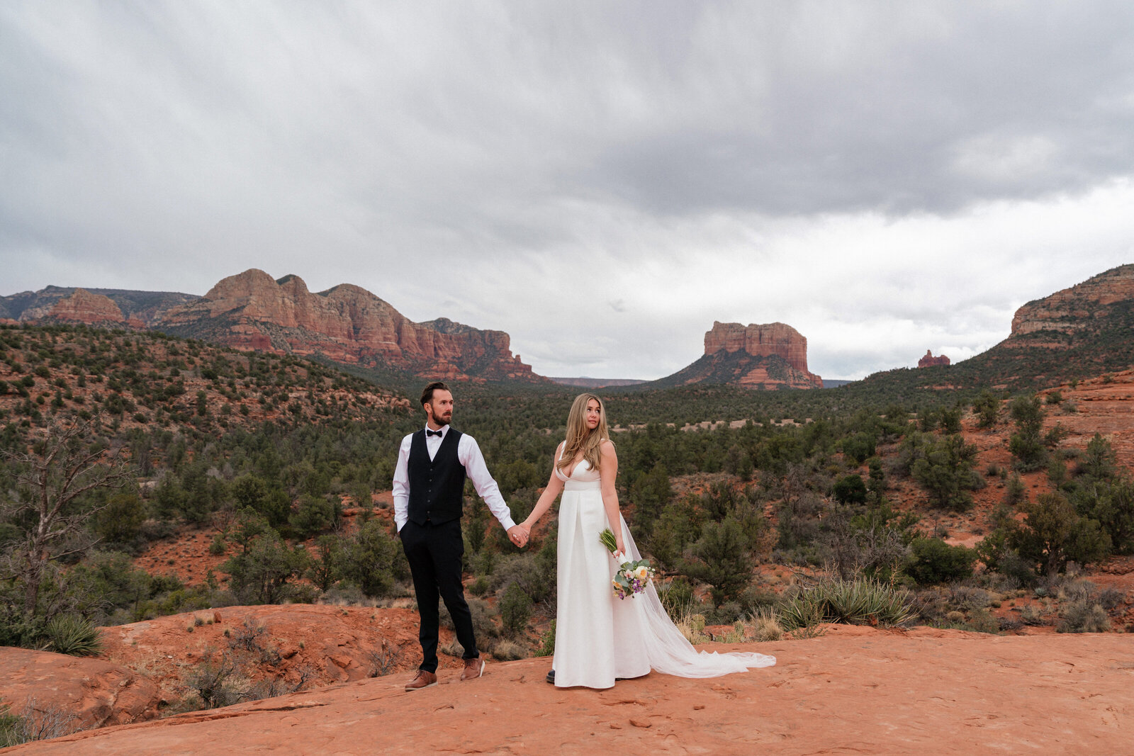 Bride and groom holding hands on a red rock overlook during their elopement at Cathedral Rock in Sedona, Arizona, with dramatic desert scenery and cloudy skies surrounding them.