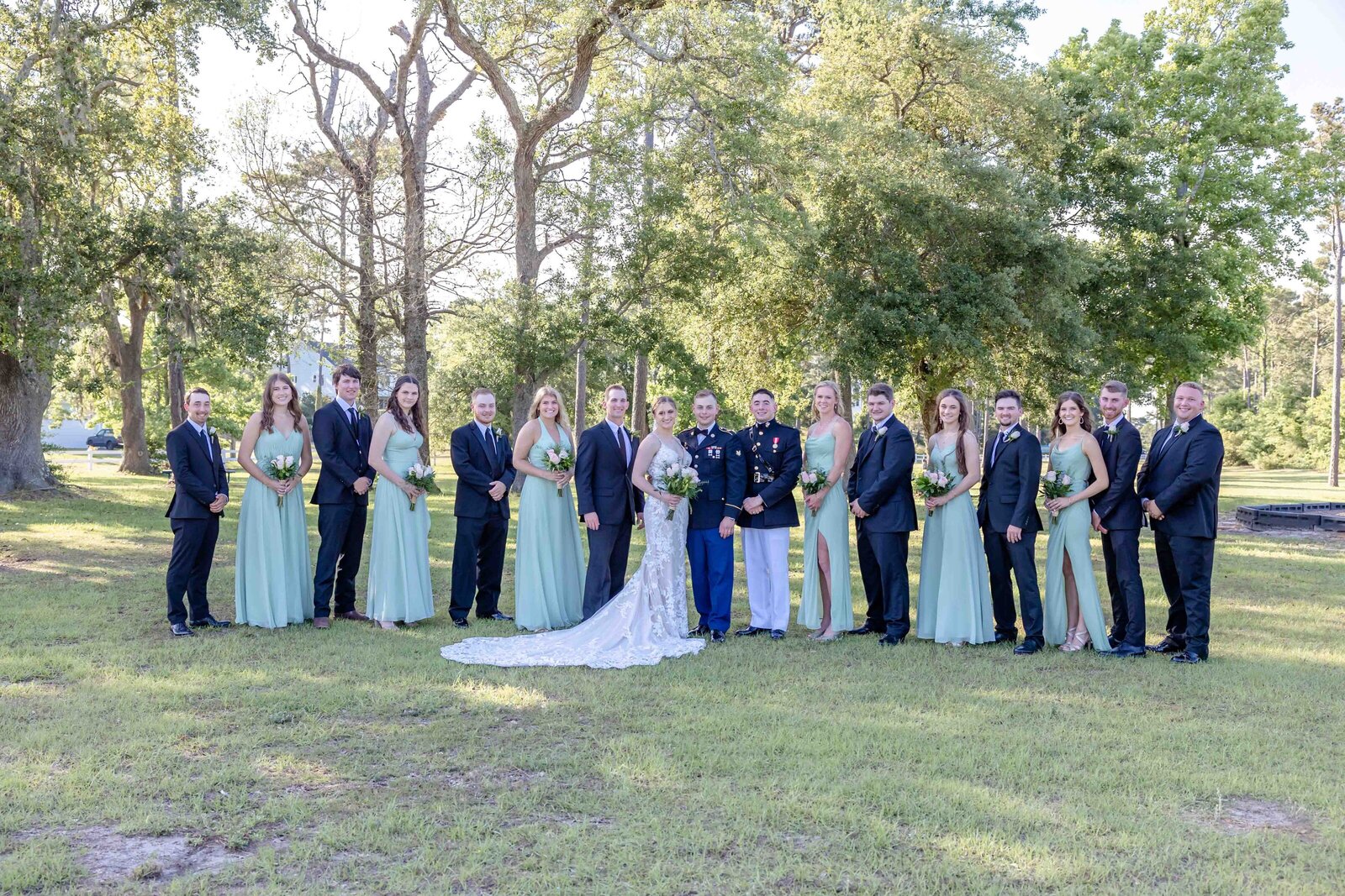 The bride and groom pose with their large wedding party dressed in sage green and navy attire on a sunny lawn, captured during a Oak Island, NC wedding, showcasing elegant group celebration.