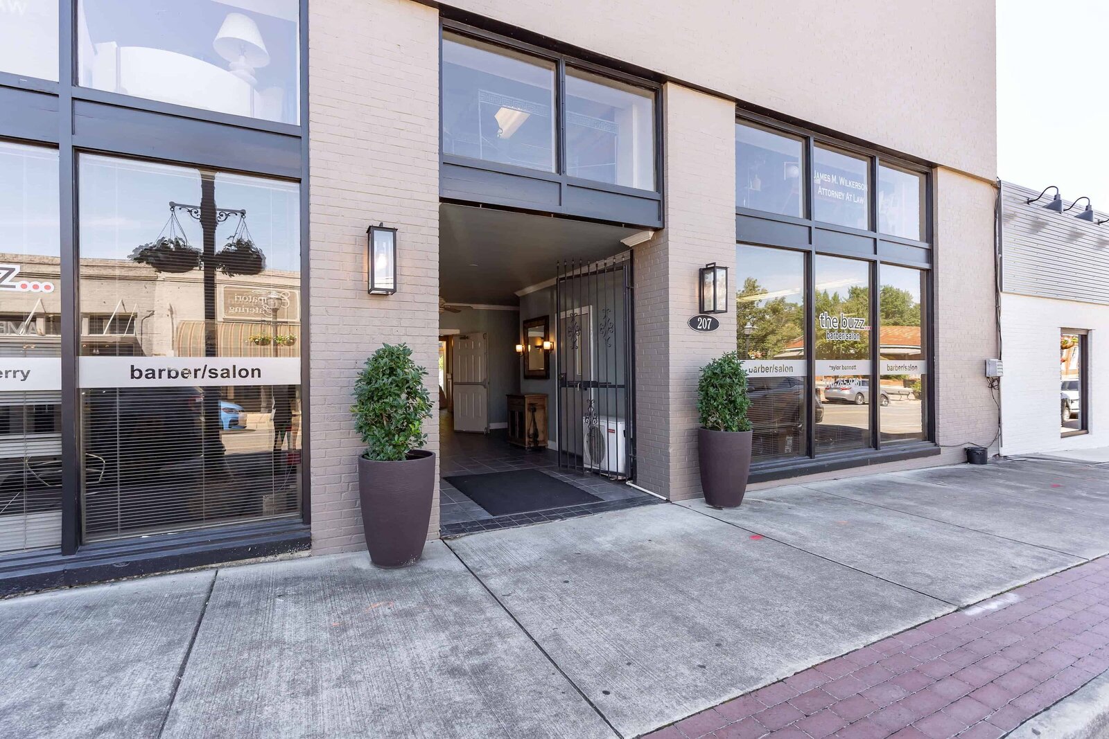 Street-level entrance to The Norton Building in Ruston, Louisiana with large windows, two potted plants by the open doorway. The sidewalk and reflections of trees and cars are visible.