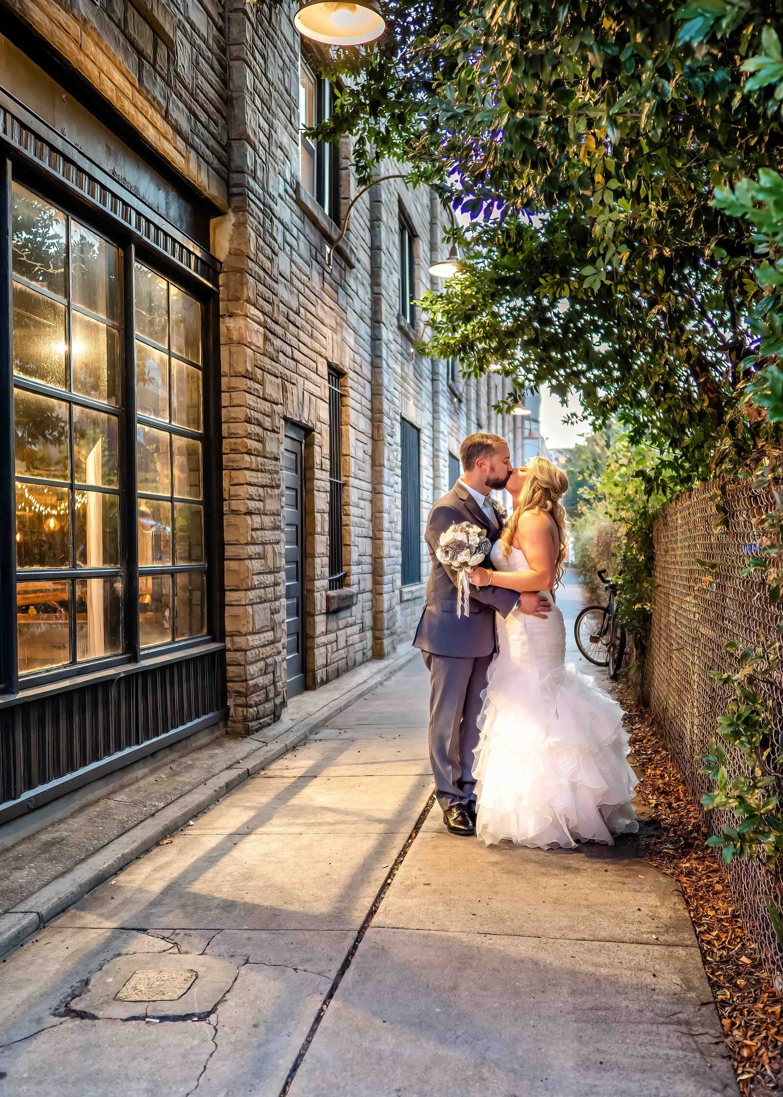 The bride and groom share a romantic kiss in a charming alley with string lights and greenery, captured during an Asheville, NC wedding, blending urban charm and romantic ambiance.