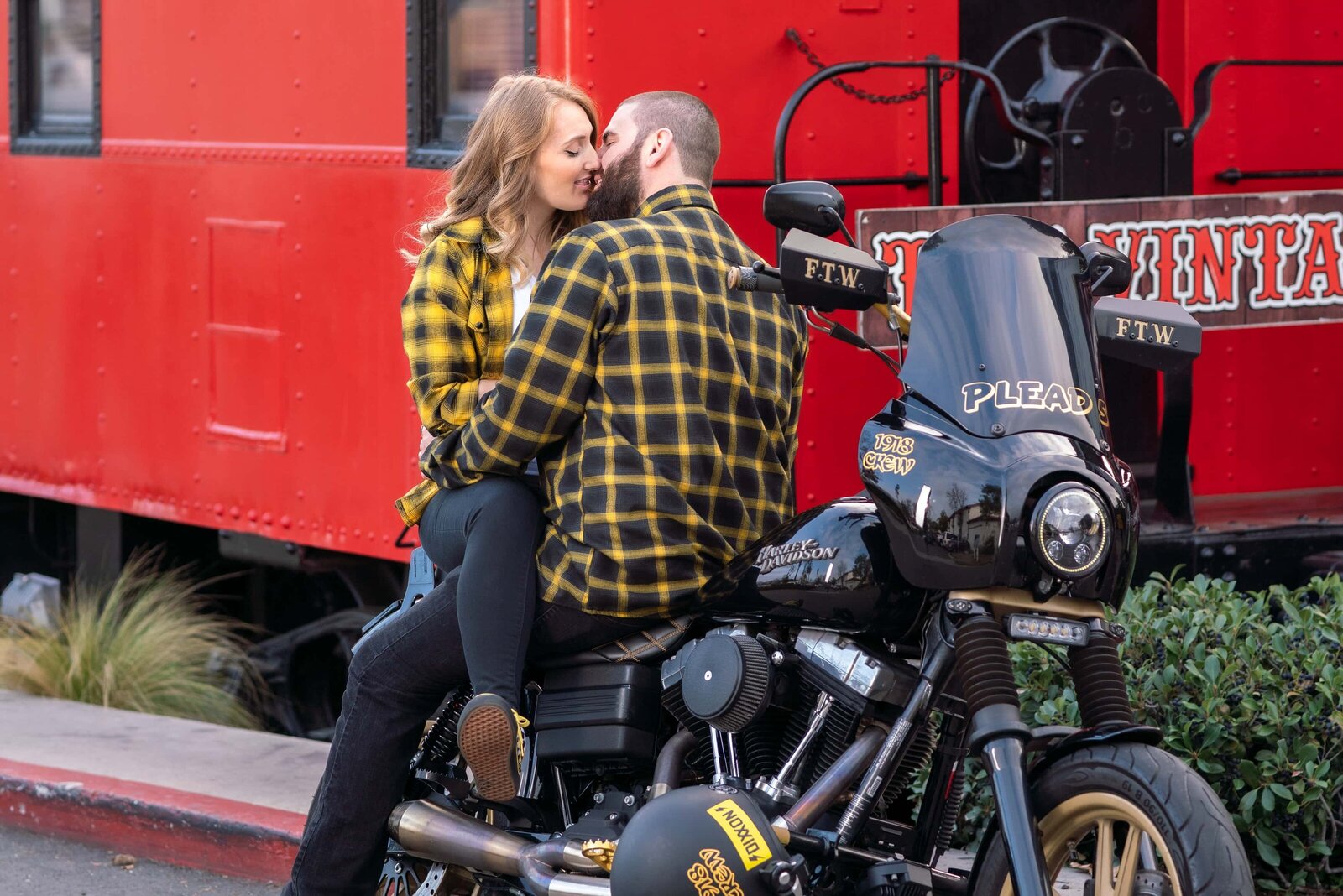 Man sitting facing blond woman. Both in yellow checkered shirt on Harley mortorcycle. Red train in background.
