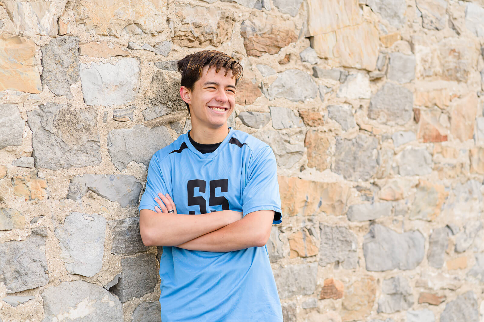 A high school senior boy wearing a blue volleyball jersey stands with his arms folded in front of a light colored stone wall in Lehi. Captured by Salt Lake City Senior Photographer Melissa Woodruff Photography