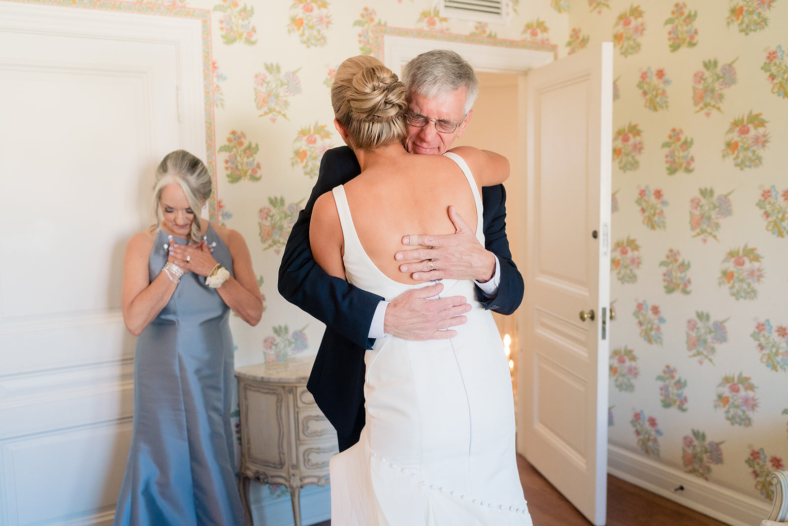 A bride and her father are hugging with the mother emotional in the background in the bridal suite at Darlington House.