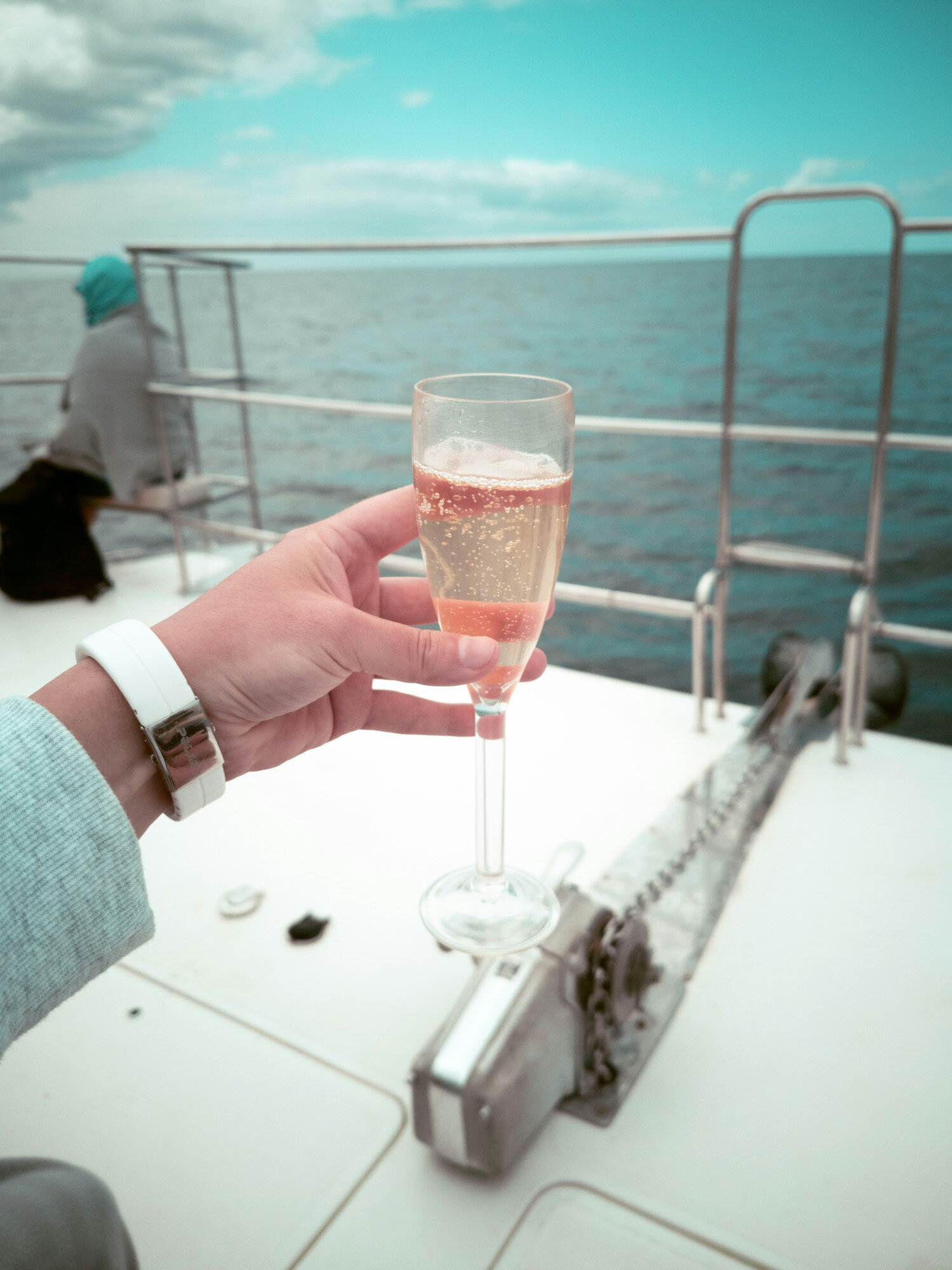 person holding a glass of champagne on the deck of a boat, with the ocean in the background and another person sits near the railing, facing the sea