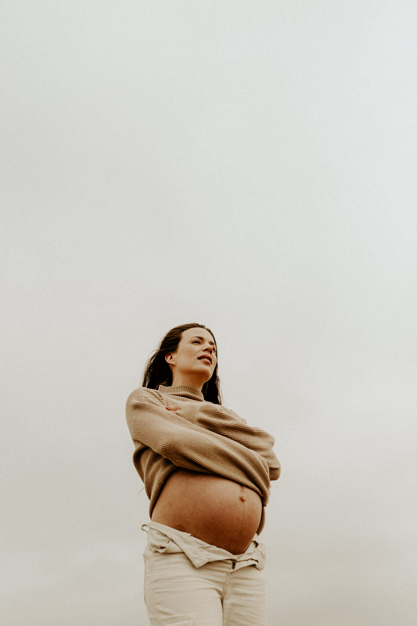en gravid kvinna i byxa och kofta håller om sig med stormiga moln i bakgrunden