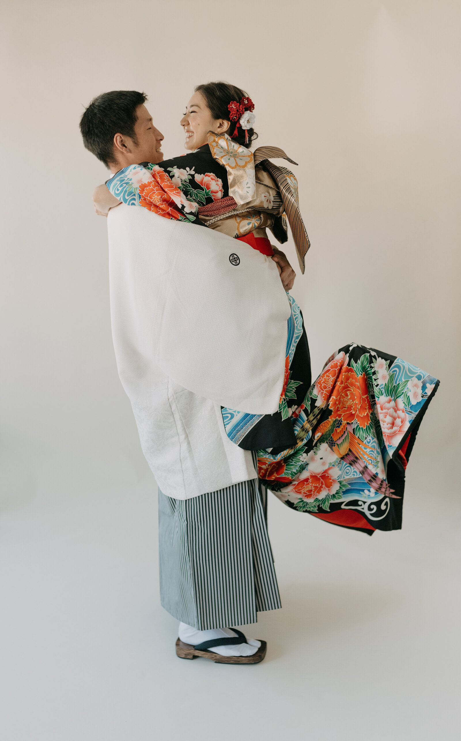 Groom lifting bride dressed in black wedding kimono rental with vibrant peony pattern for a joyful studio portrait, Los Angeles California | 黒引き振袖レンタル 花嫁を抱き上げる新郎 幸せあふれるスタジオウェディングフォト