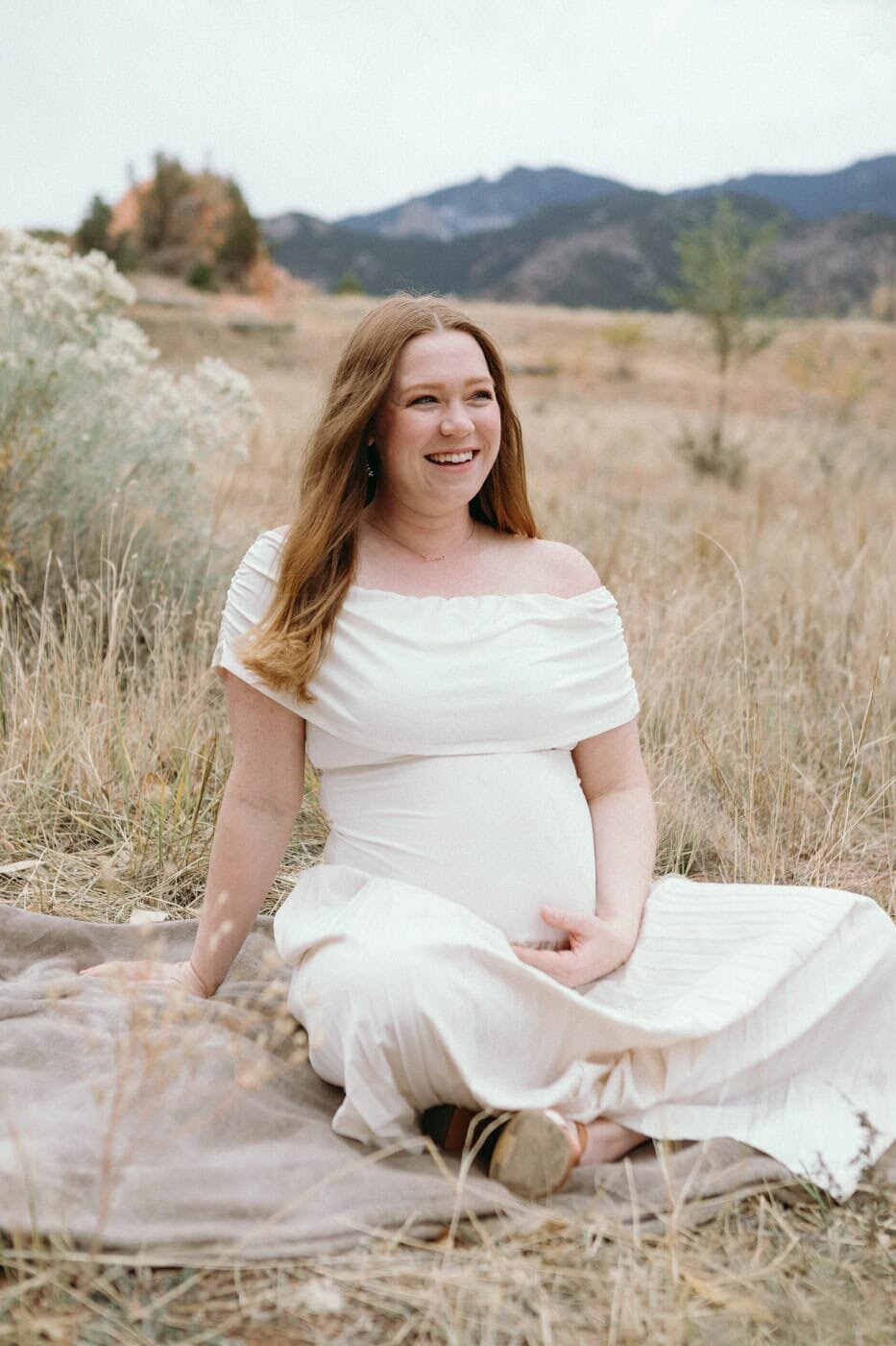A pregant woman sits in yellow grass holding her belly and smiling.