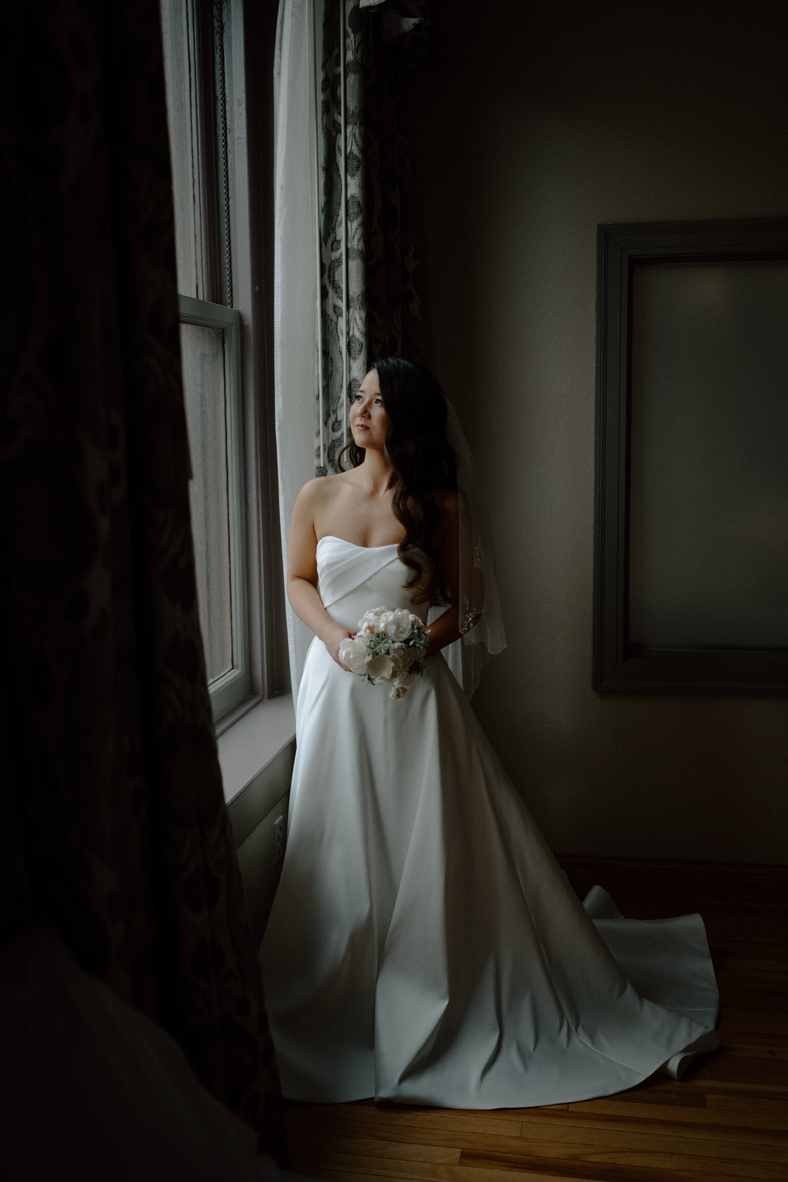 Bride standing by a window in an elegant wedding gown with polished, romantic hair styled by Promise Artistry.