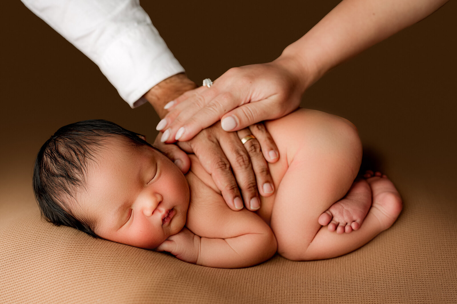 A newborn baby sleeps peacefully on a brown surface, curled up with dark hair and a calm expression. Two adult hands gently rest on the babys back, offering protection and care—captured beautifully by a Charlotte newborn photography studio.