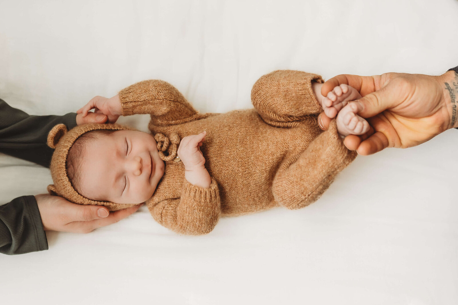 Newborn baby boy in a fuzzy bear romper with mom holding head and dad holding feet