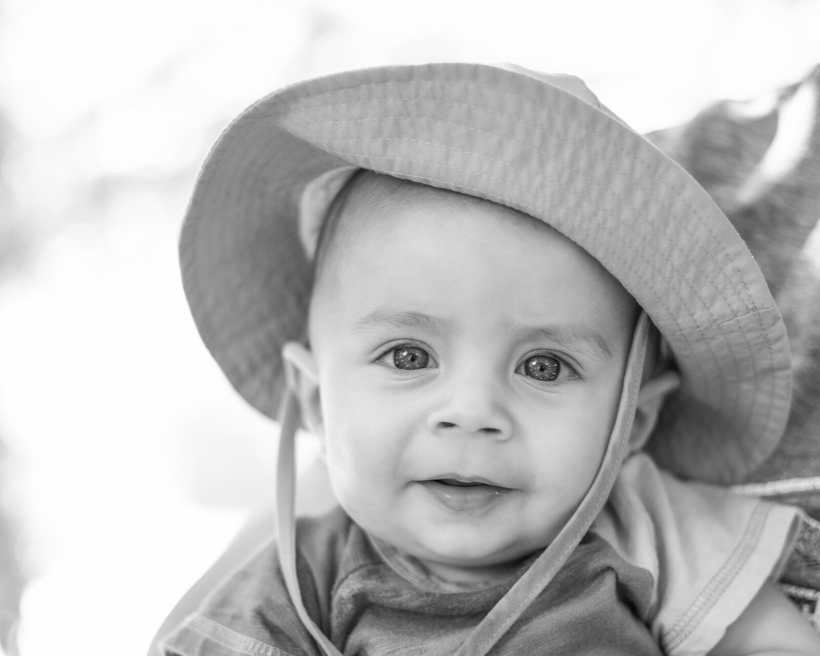 a black and white photo of a baby wearing a hat, one piece outfit, and looking at the camera with a soft smile