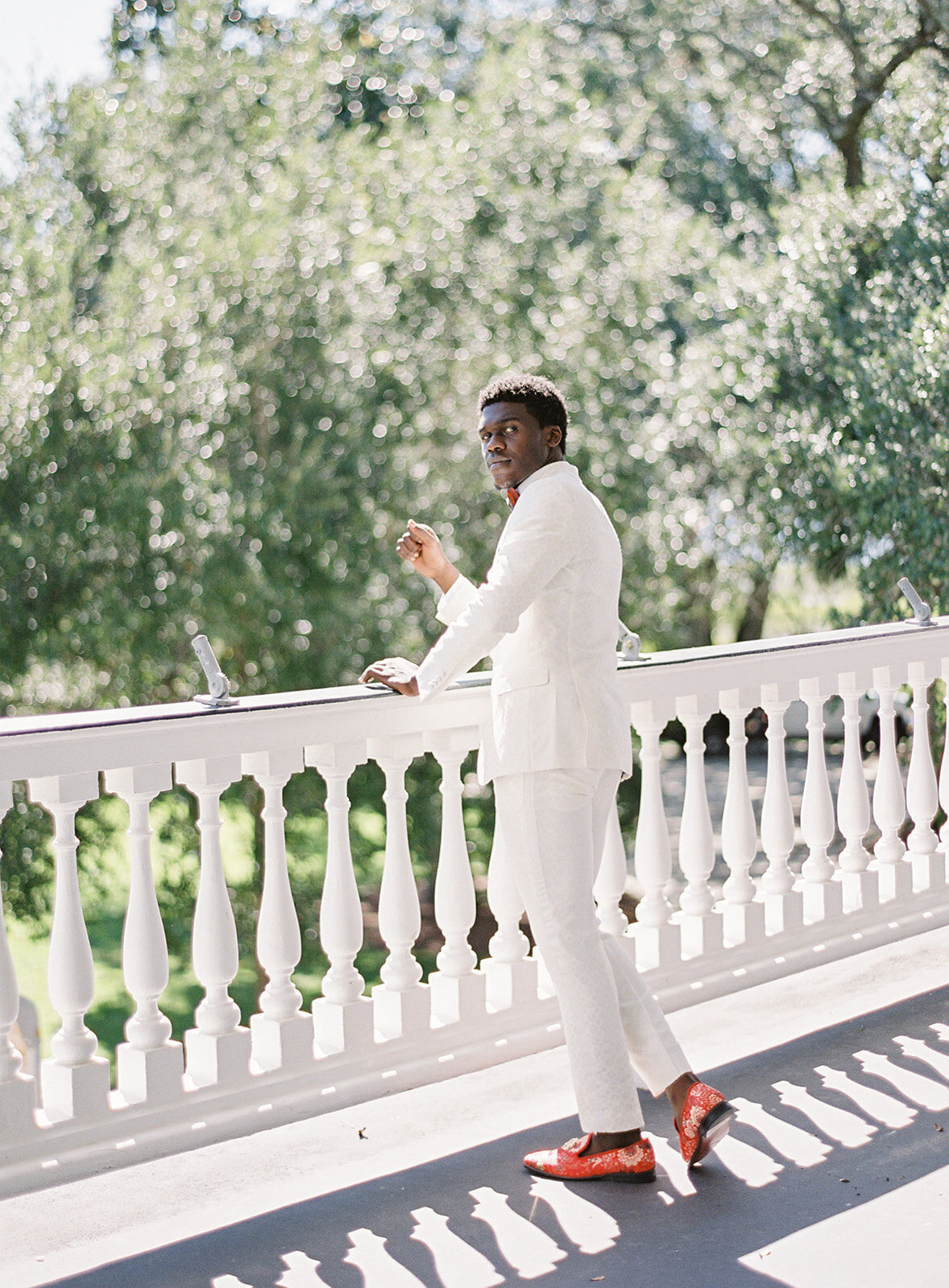 Groom in ivory tuxedo with gold detailing and red shoes standing by a white railing of balcony. His left hand is on the railing and he is looking in to the camera. Photographed by wedding photographers in Charleston Amy Mulder Photography.