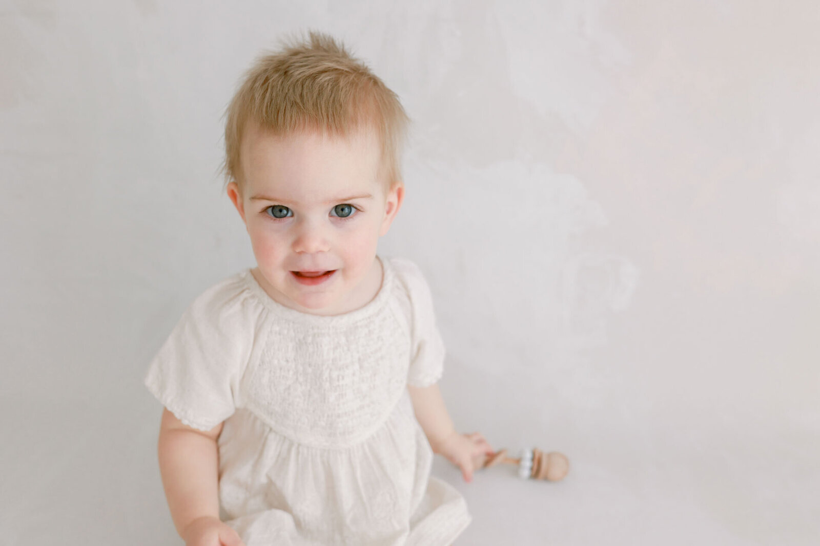 Ein Babyfoto in zarten weiß Tönen. Blondes Baby mit blauen Augen im weißen  Kleidchen sitzt vor einem hellen Hintergrund und sieht interessiert in die Kamera.