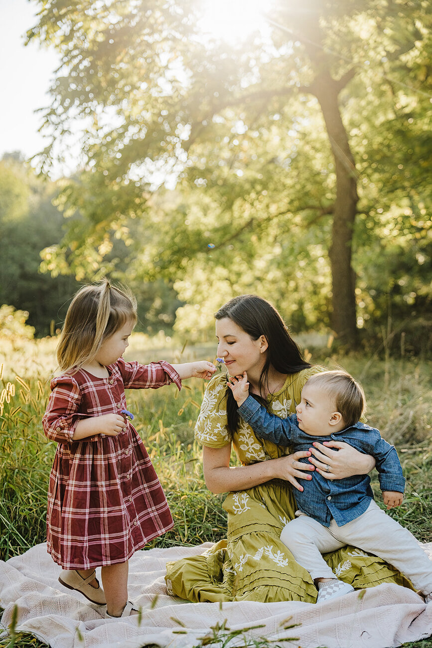 mother sits on blanket with baby son and daughter and smells flowers at sunset