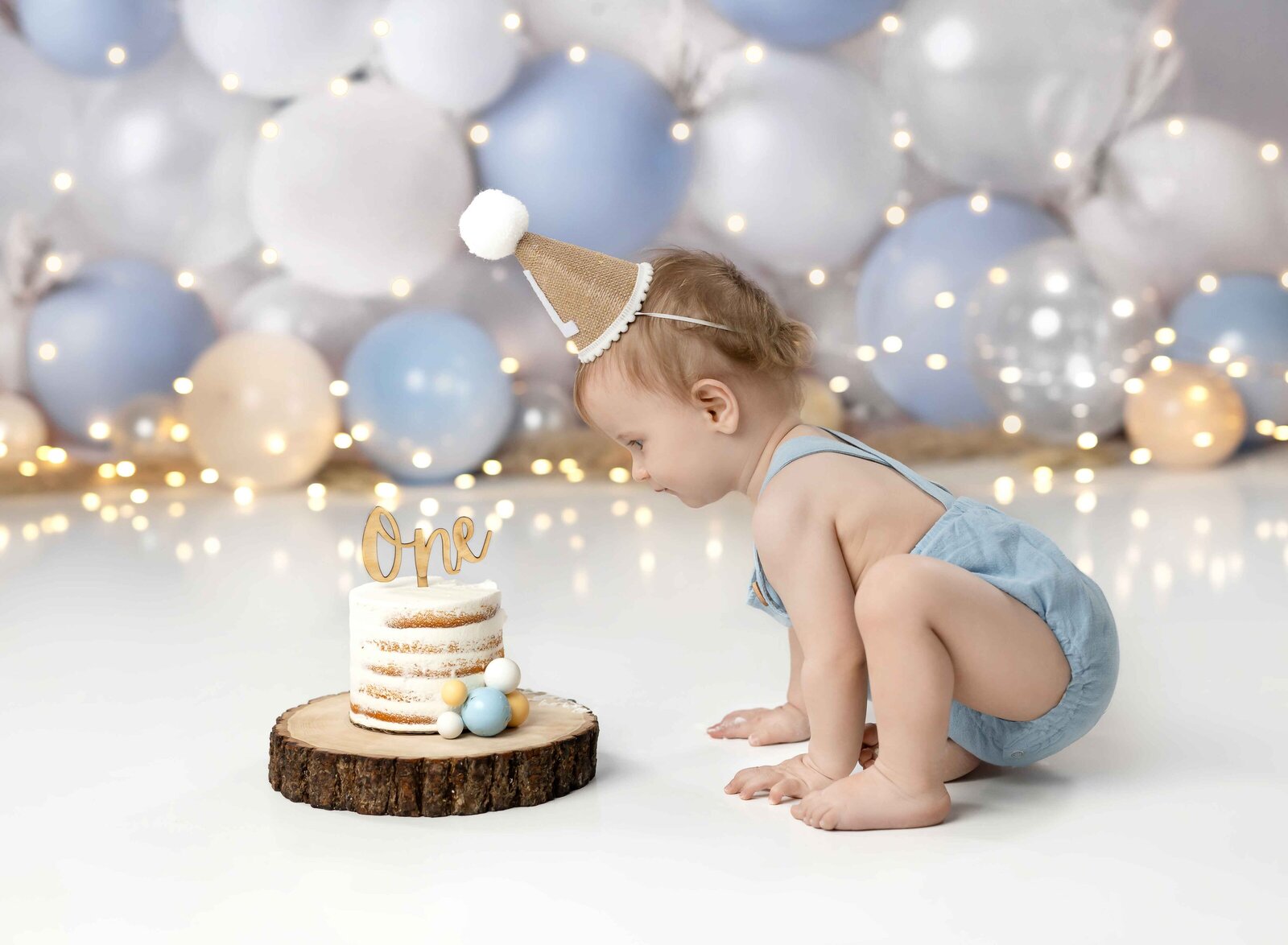 Baby boy in blue romper, crawling towards birthday cake and looking at it.