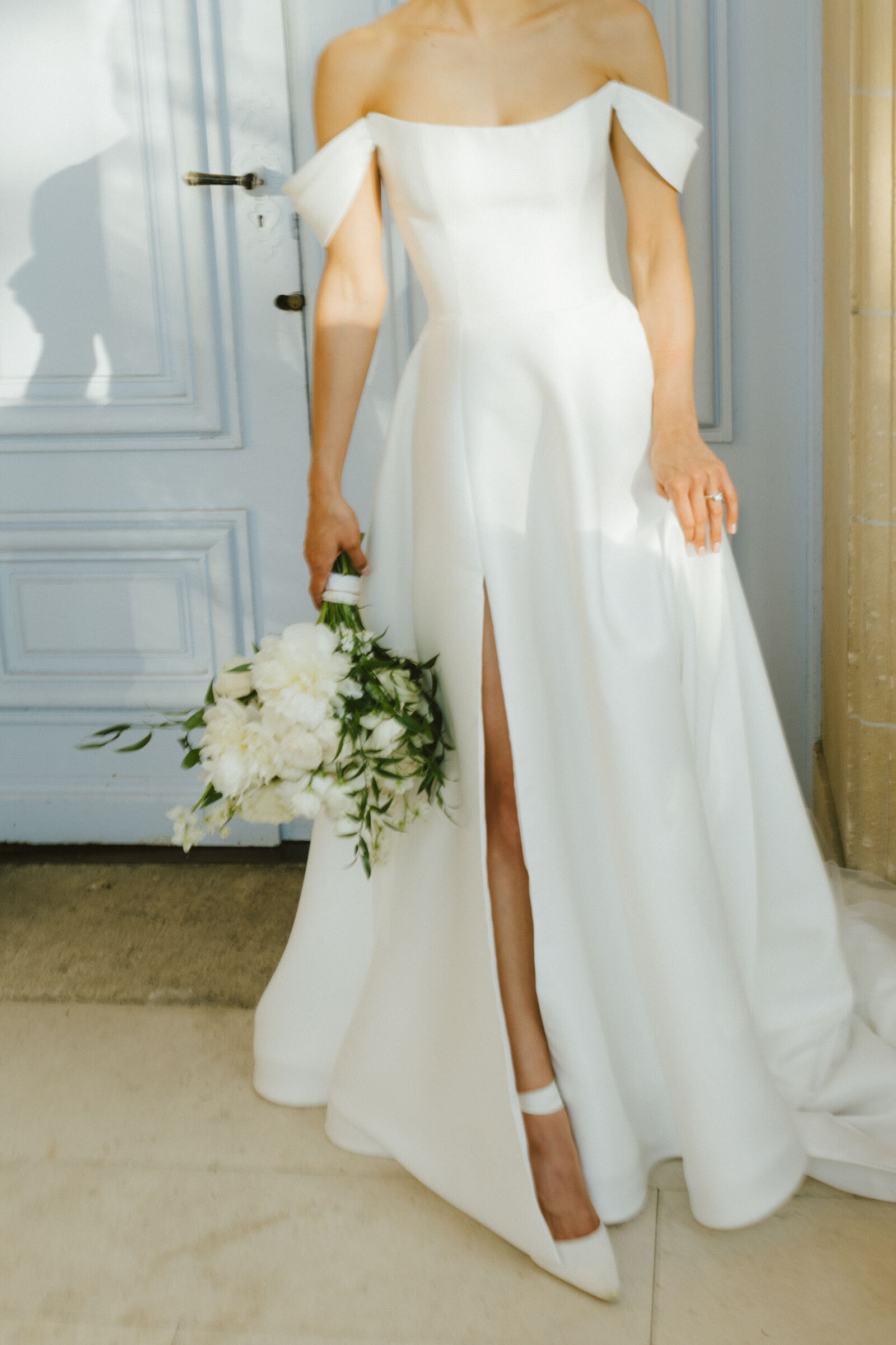 A bride shows off her wedding dress and bouquet in front of blue French doors in Paris.