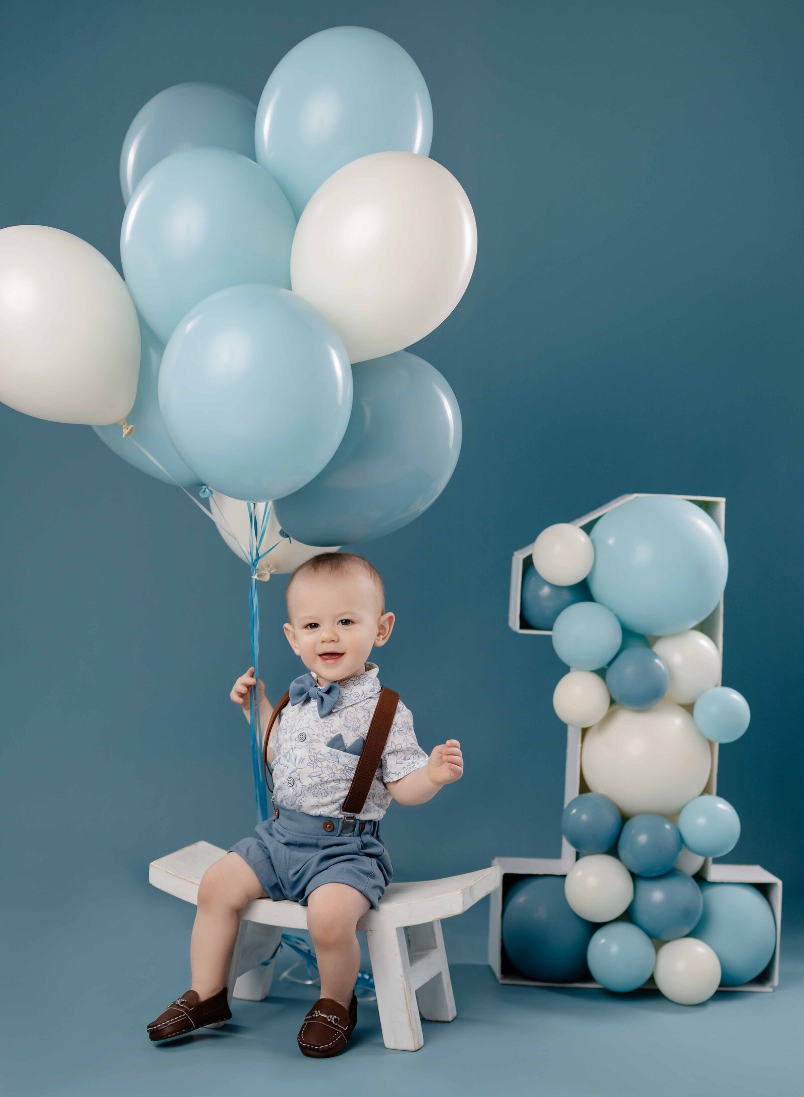 Baby sitting on stool, holding balloons and smiling at camera.
