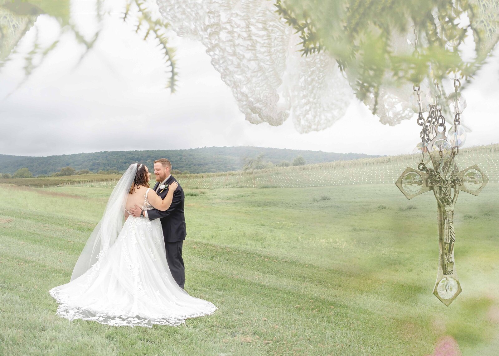 The bride and groom share a romantic moment in a mountain meadow with a hanging cross and delicate lace details in the foreground, captured during a Boone, NC wedding, blending rustic charm and spiritual elegance.