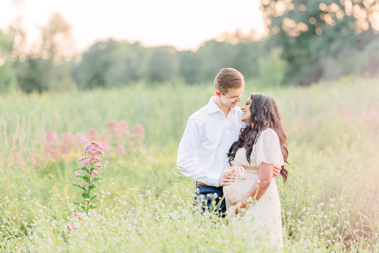 Man and his pregnant wife smile at each other with his arm around her in a field of tall wild flowers