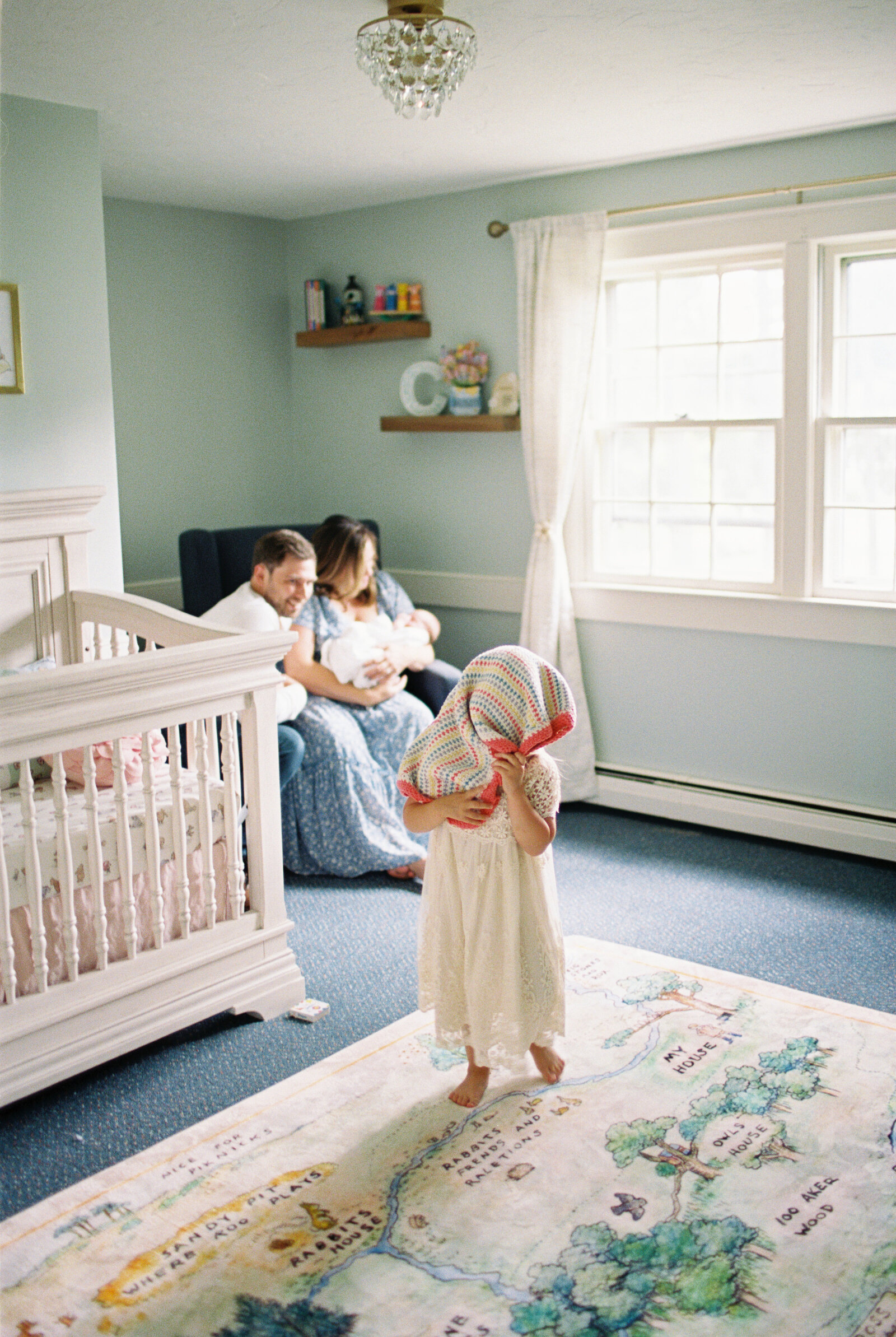 A young toddler is standing in her new baby sister's room while playfully putting a blanket on her head