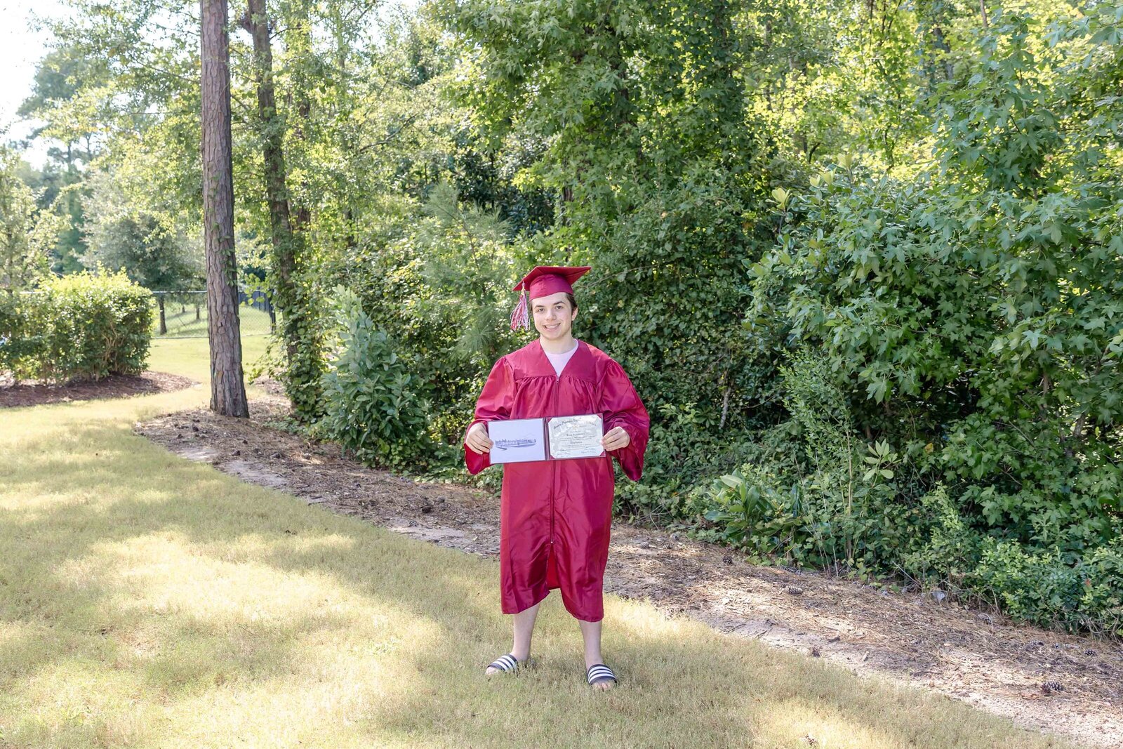  A high school senior in a red graduation gown proudly holding a diploma in a park setting in Wilmington, North Carolina. This celebratory image captures the joy of graduation, perfect for capturing meaningful senior portraits.