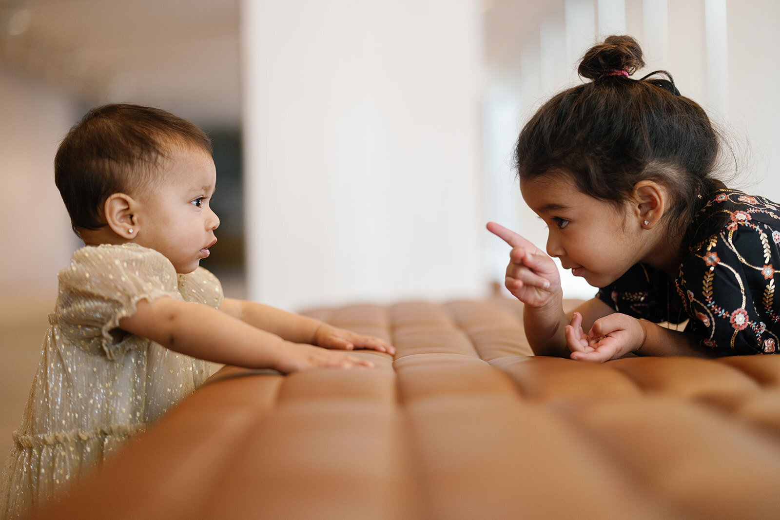 A lively moment between two young sisters on a leather bench, as one playfully points at the other, capturing their unique bond and energy.