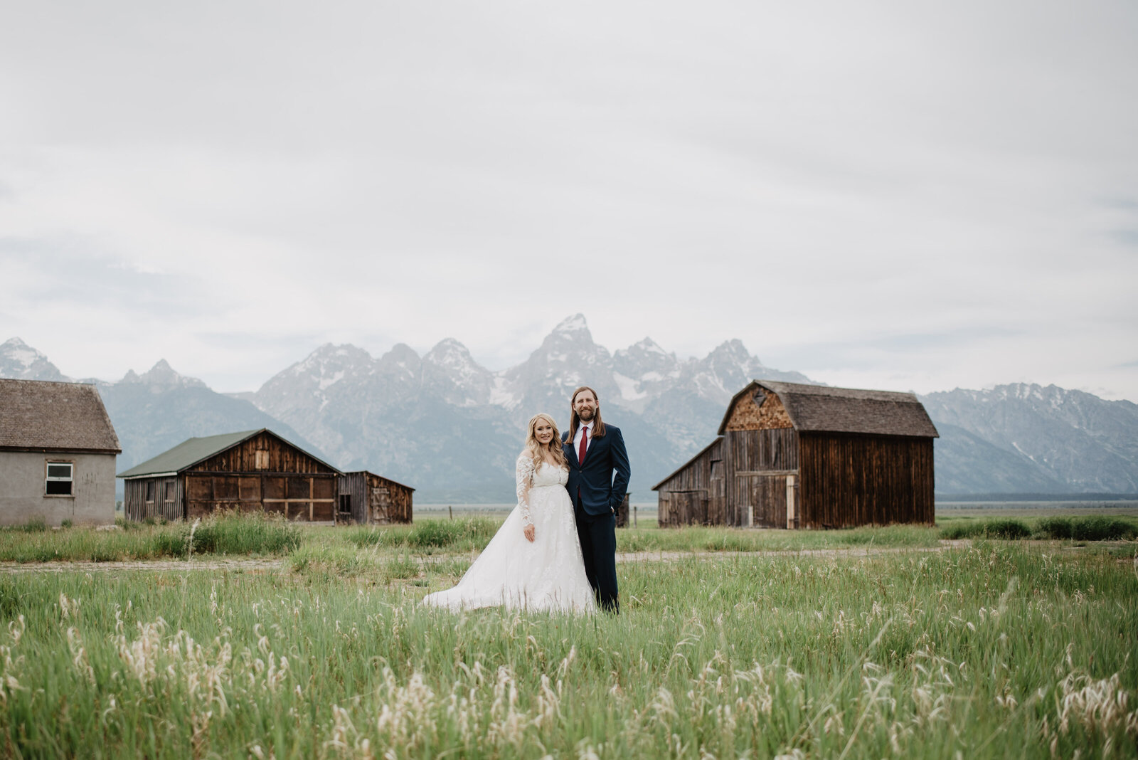 jackson hole wedding  photos with bride adn groom standing in a open field together and posing as they stand together and smile with the grand tetons behind them past a few barns