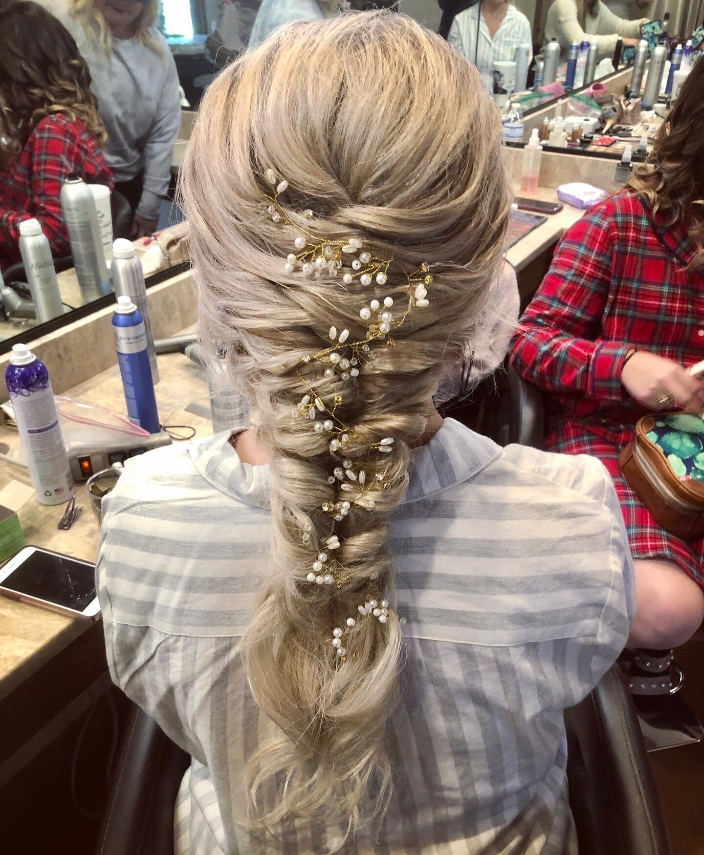 In a salon setting, this blonde braided hairstyle adorned with pearl and floral accessories offers a glimpse into the ultimate bridal beauty experience in Texas, where styling products surround you for that flawless wedding beauty consultation.