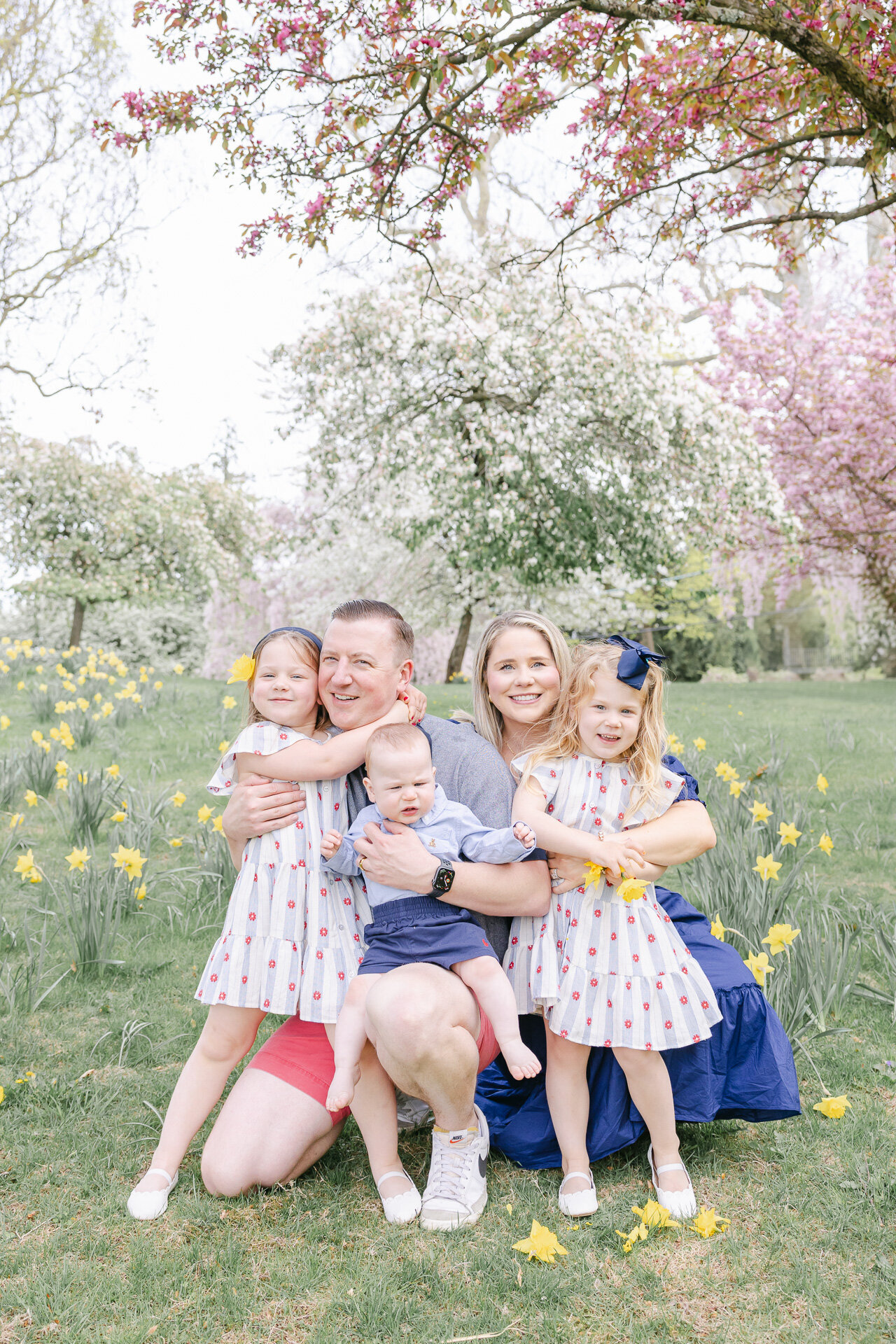 A family of five poses joyfully in a blooming Connecticut field during a springtime photoshoot. Surrounded by daffodils and flowering trees, the parents kneel with their three young children—twin girls in matching polka dot dresses and a baby boy in blue. Their coordinated outfits and genuine smiles capture a picture-perfect seasonal moment.
