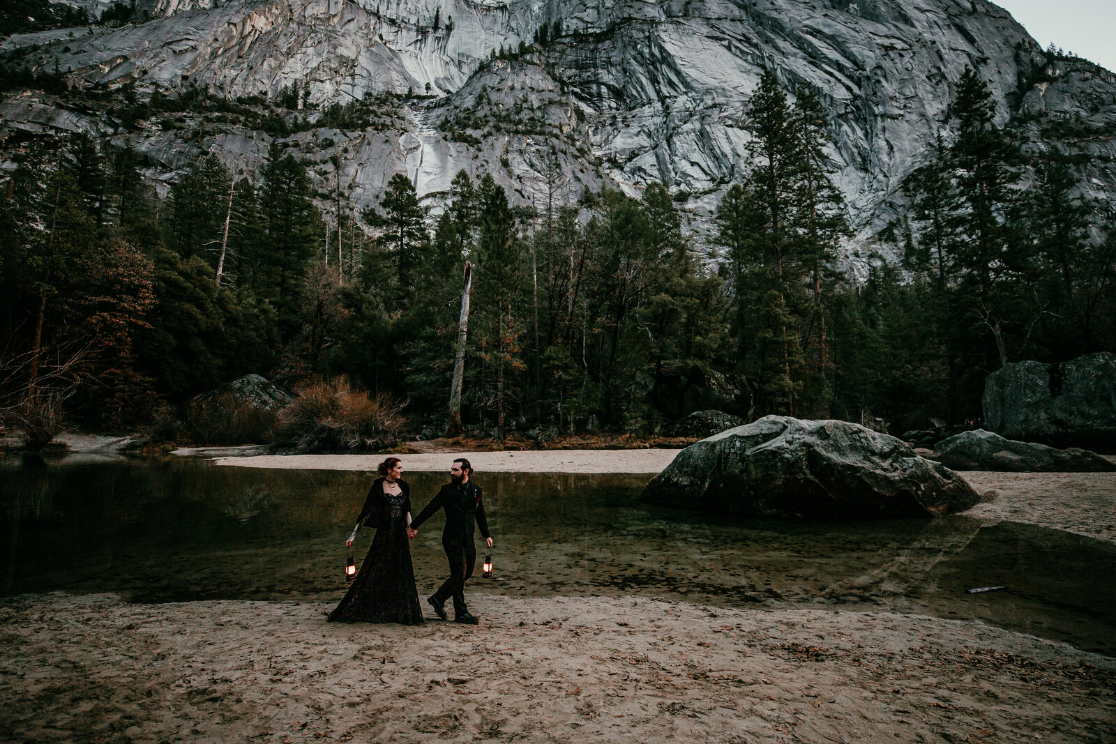 A couple dressed in all black walks hand in hand along the water’s edge at Mirror Lake in Yosemite, lanterns glowing as they embrace the moody, adventurous vibe of their elopement.