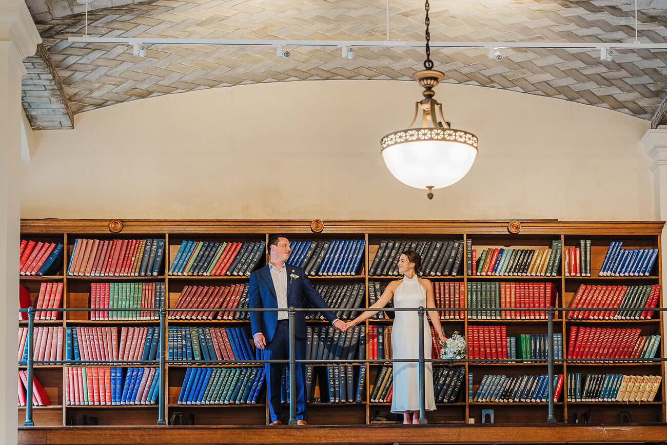 wedding couple stands in boston public library holding hands in front of book stacks
