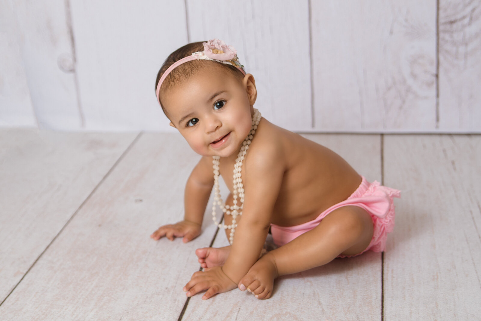 Milestone Photographer, a baby wears a necklace of pearls and sits up on wooden floors