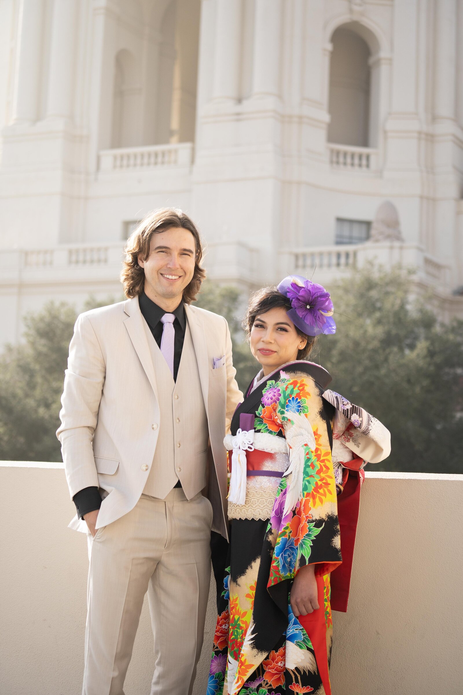 Bride in black wedding kimono rental with vivid multicolor floral design and groom in beige suit posing in front of the grand architecture of Pasadena City Hall, Pasadena California | パサデナ市庁舎前で黒地に金彩花柄の振袖をレンタルした花嫁とベージュスーツの新郎が並ぶ着物ウェディングフォト