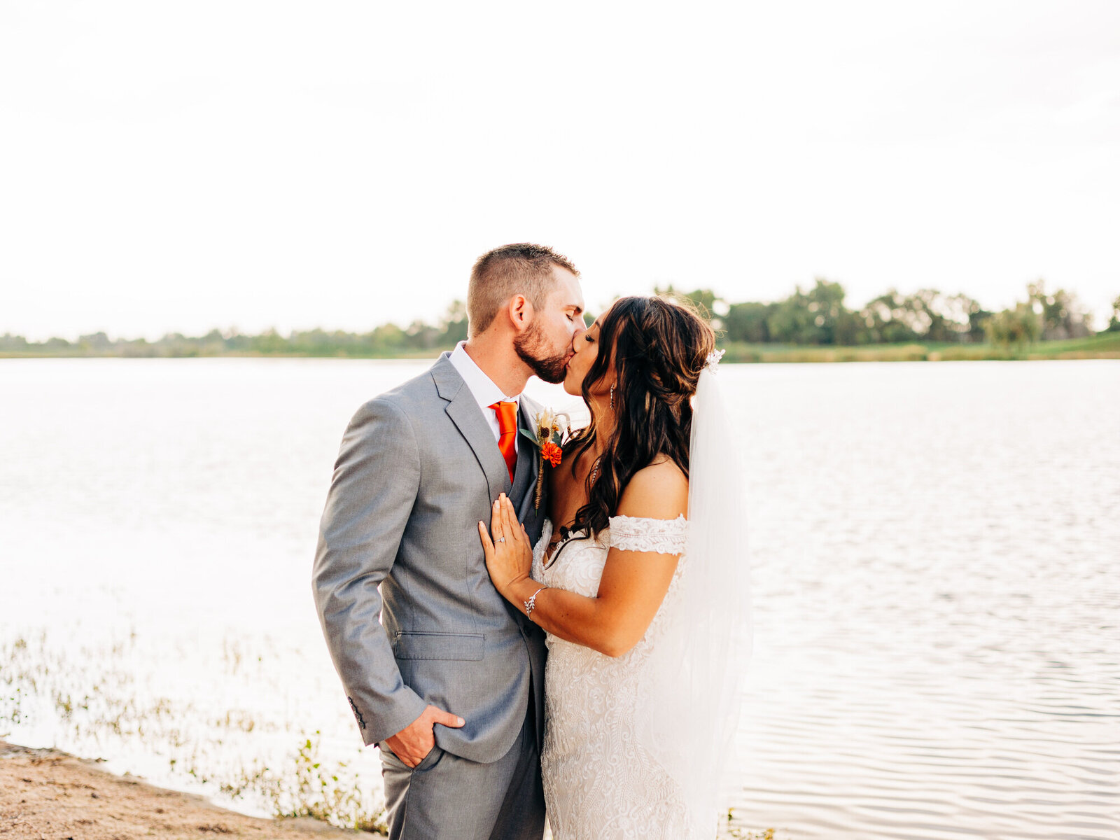 A bride and groom share a kiss by a lake. Captured by an Eau Claire Wedding Photographer, the groom wears a light gray suit with an orange tie while the bride stuns in an off-the-shoulder white gown. Trees and water frame this romantic scene.