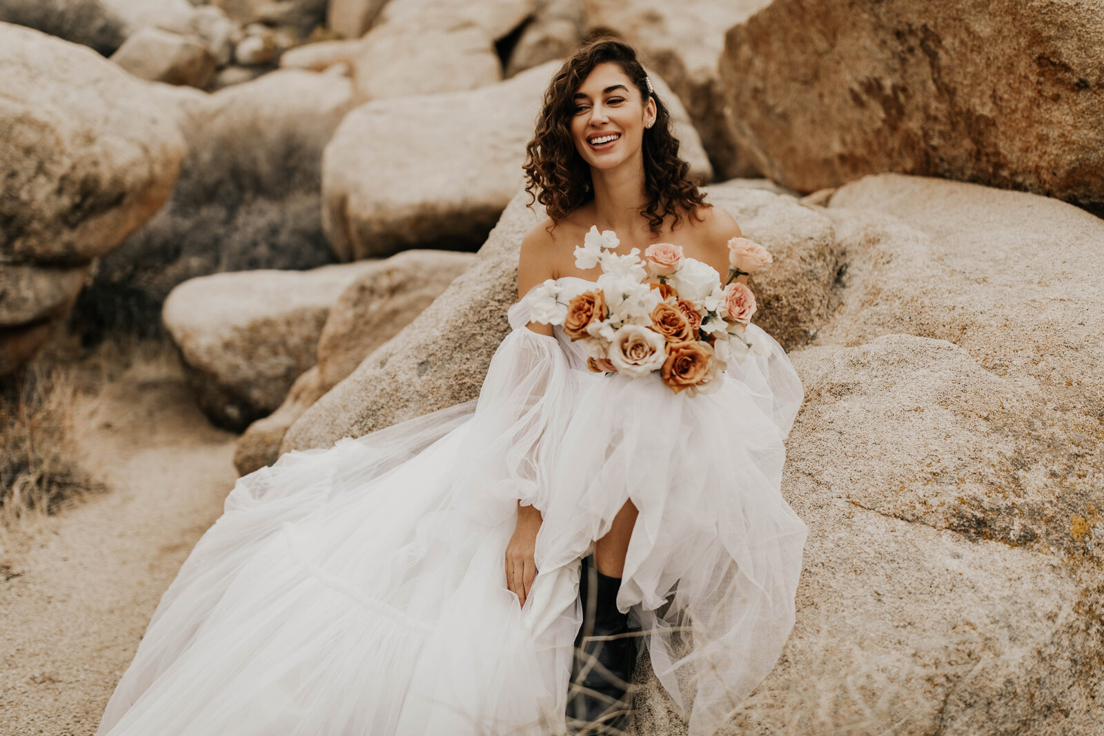 Bride holding bouquet smiling sitting on desert boulders