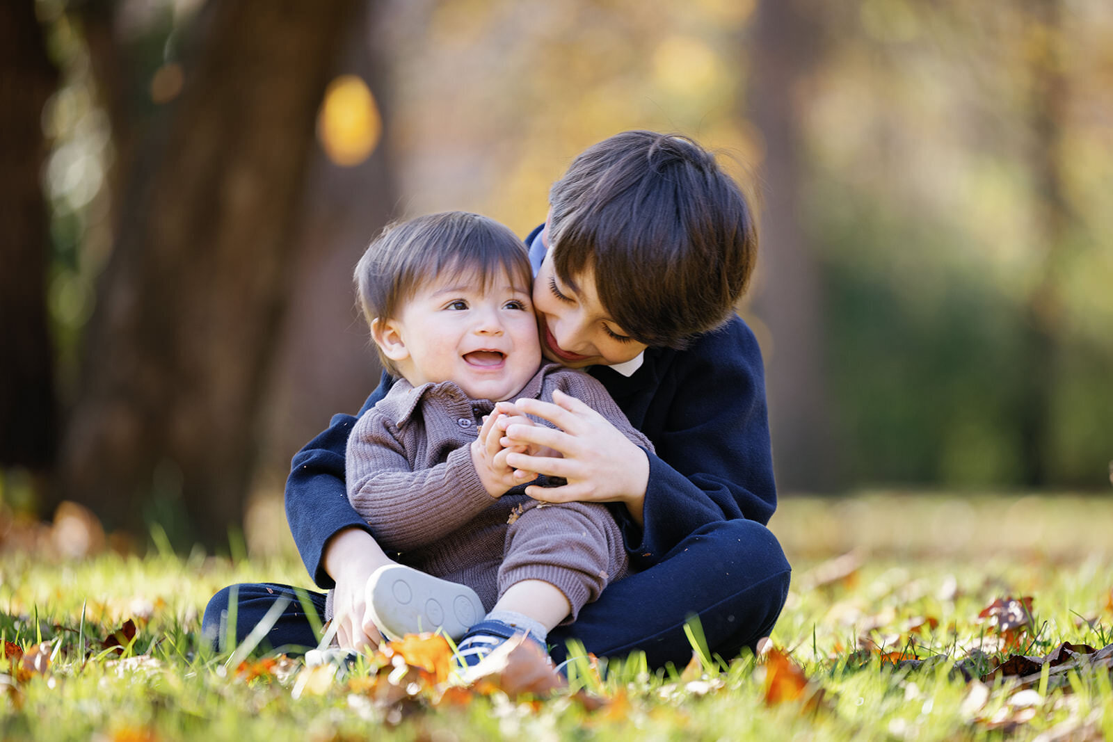 A heartfelt outdoor moment of two brothers sitting on grass, with the older one lovingly embracing the younger, surrounded by autumn leaves.