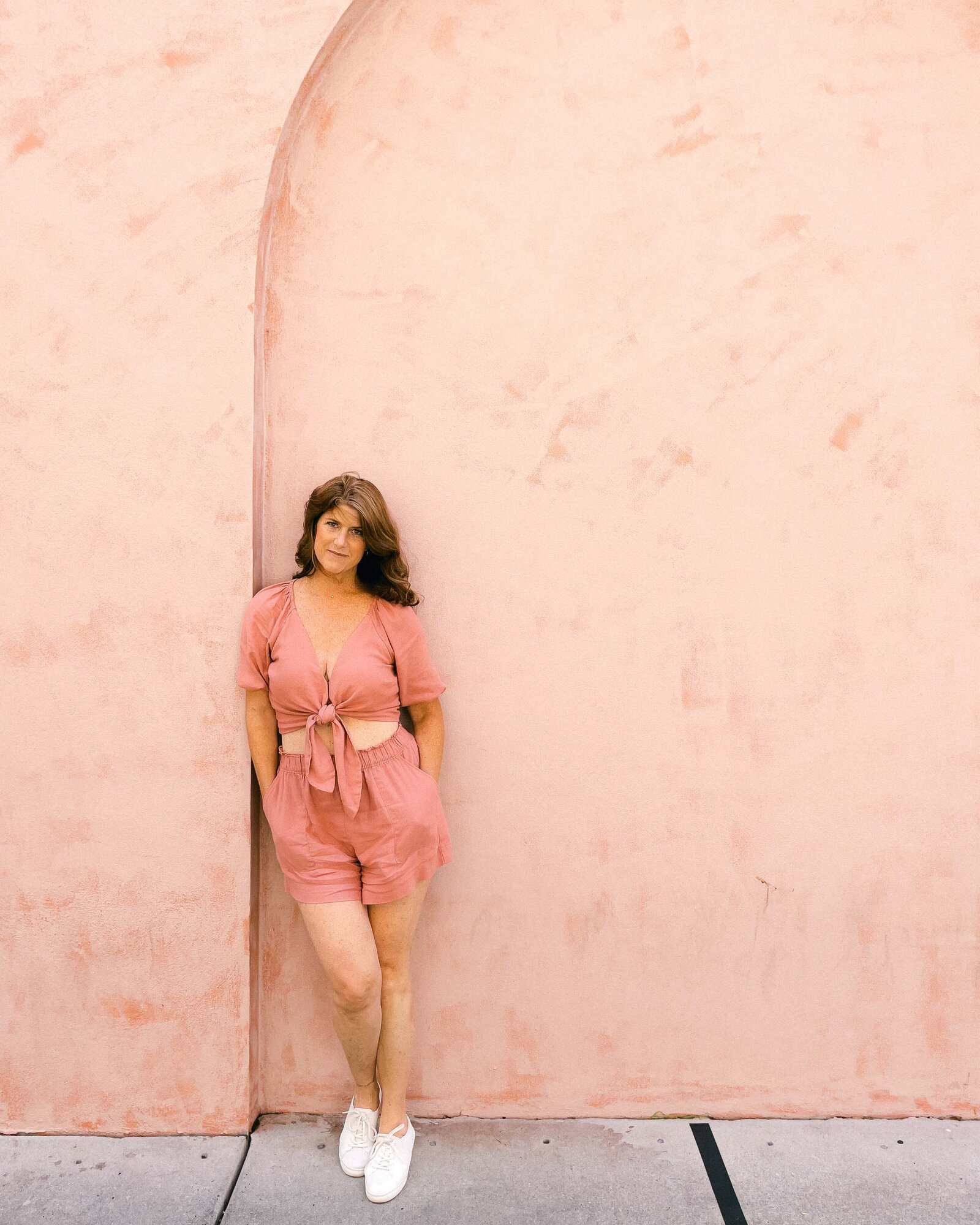 Pink wall and pink romper woman Gayle Dawn leaning up against wall for social media posing photos.