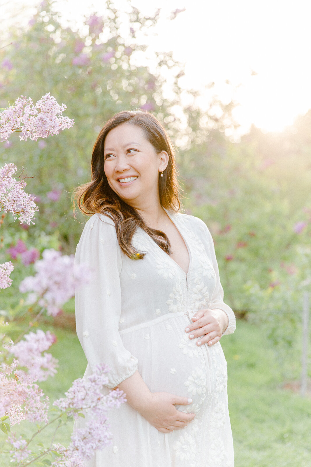A pregnant mother embracing her baby bump while standing amongst lilac blooms during photo session with Boston family photographer Corinne Isabelle at the Arnold Arboretum