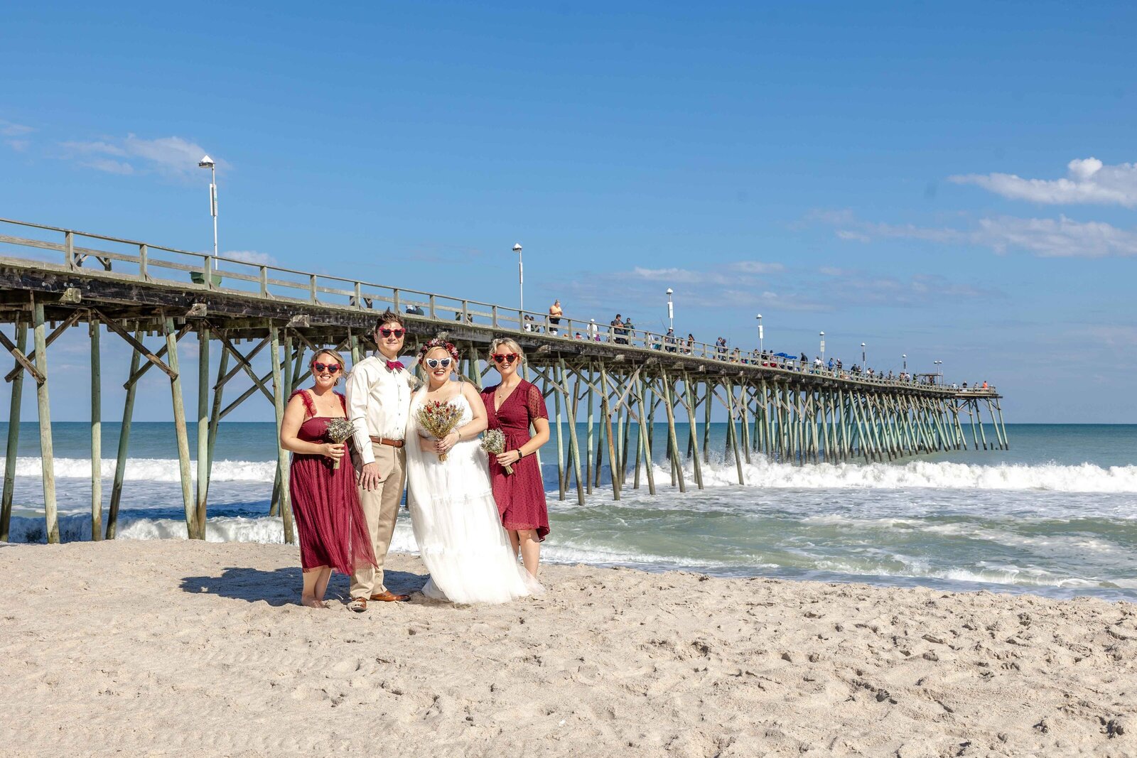 The bride and groom pose with their bridesmaids near the iconic fishing pier at Carolina Beach, NC, blending natural coastal beauty with vibrant wedding moments.