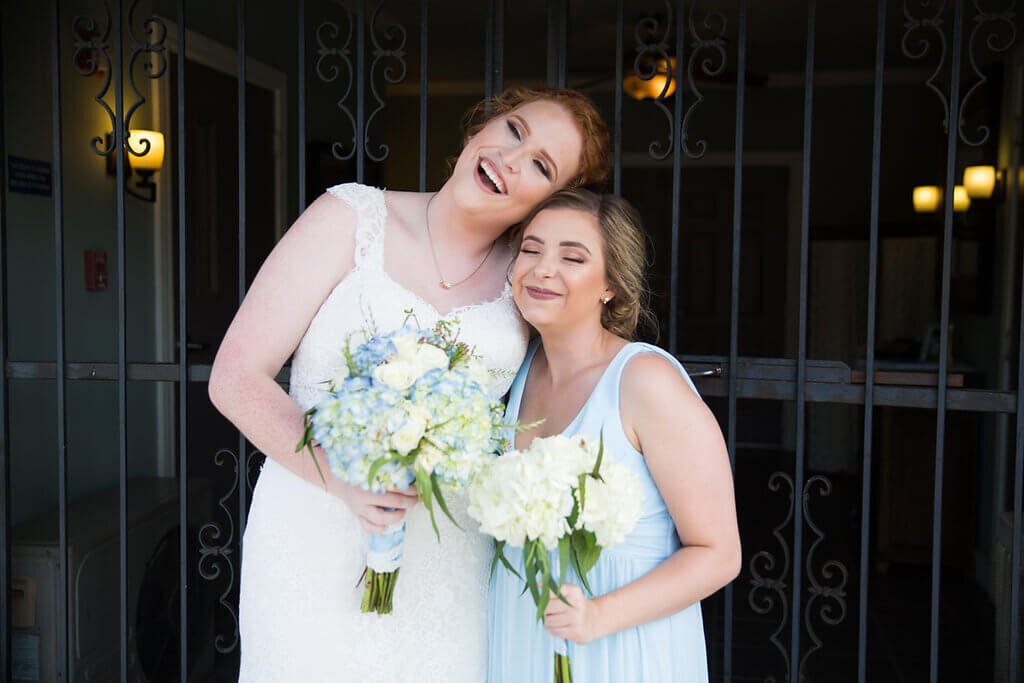 A bride and a bridesmaid wearing a light blue colored dress smile and stand close together, each holding a bouquet of white and blue flowers, in front of a decorative metal gate.