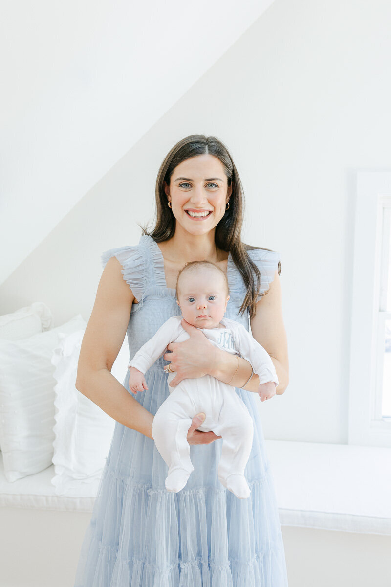 A smiling mother in a light blue dress holding her newborn baby boy in a bright, airy bedroom during an in-home newborn photography session in Fairfield County, CT, by Nicole Neira.