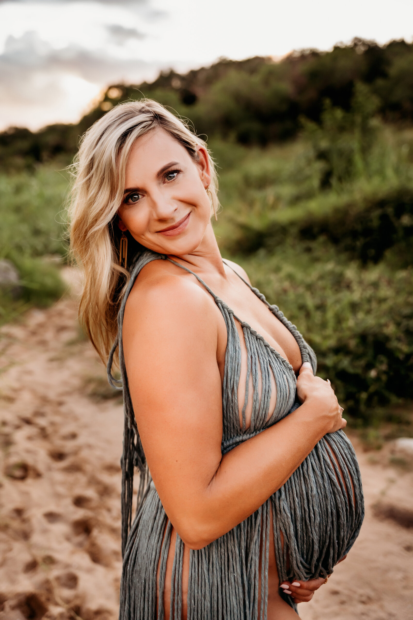 Maternity Photographer, an expectant mother wears a lacy dress and smiles in anticipation, she is at the river bank