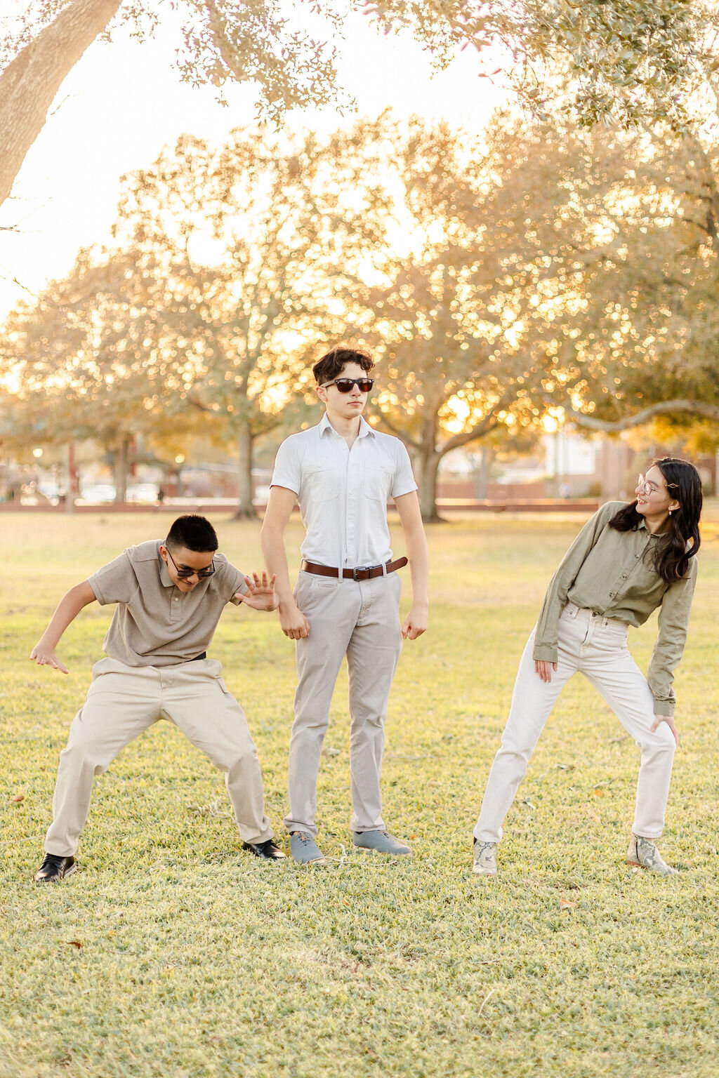 Three young adults stand in a grassy park during golden hour, with sunlight streaming through the trees behind them. The person in the middle stands upright and expressionless, holding hands with the other two. The person on the left crouches in a playful pose, while the one on the right leans toward the center with a smile. All three wear glasses and casual clothing, creating a fun and relaxed vibe.