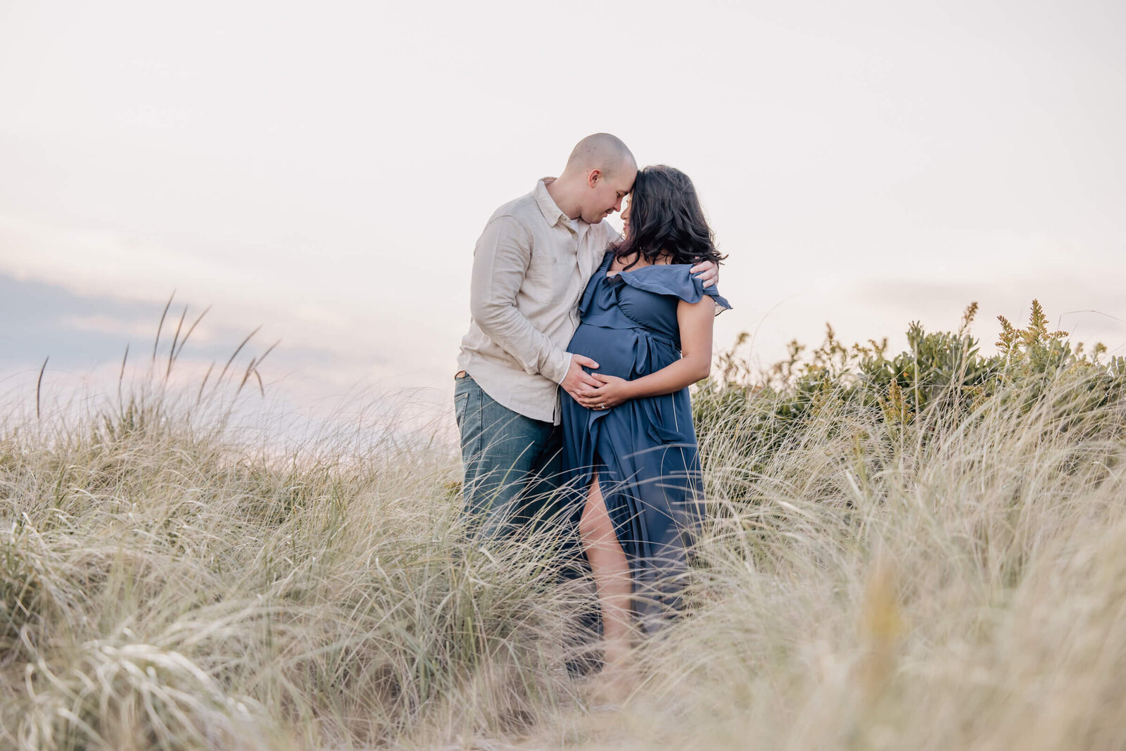 Pregnant woman and husband in tall grass near beach.