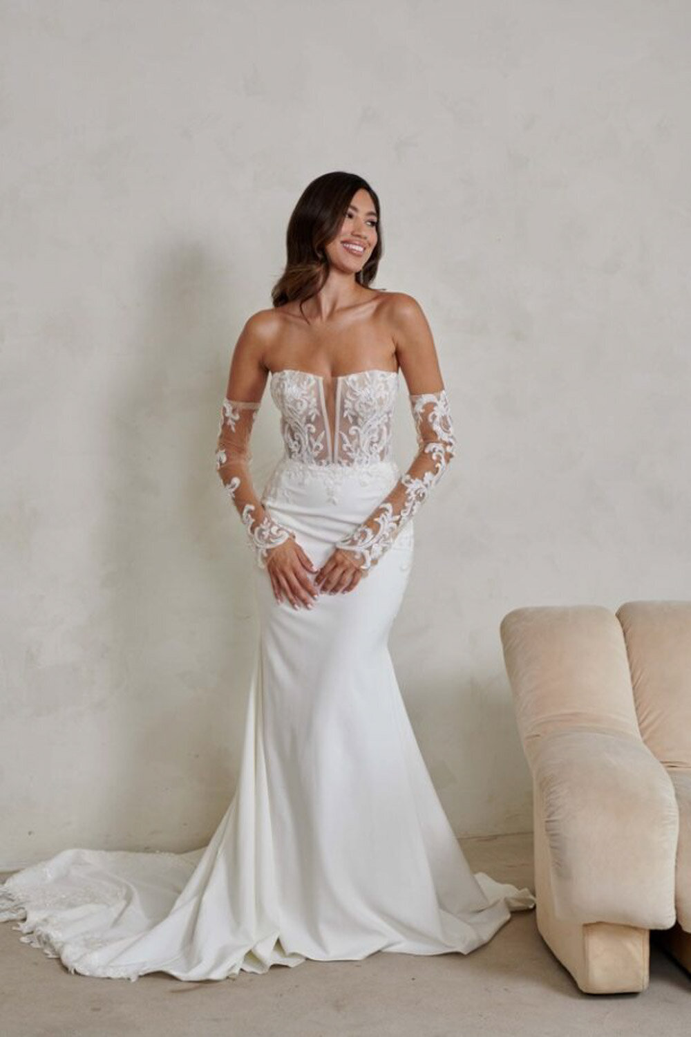 Bride in a strapless fitted gown with lace sleeves standing against a neutral wall