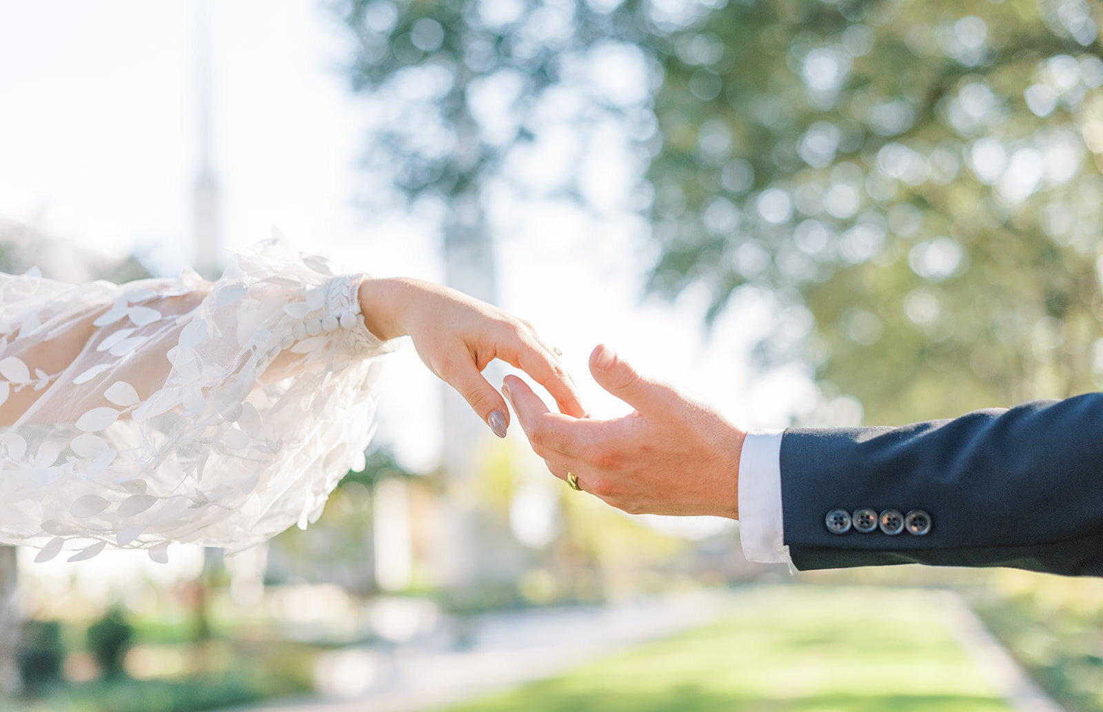 Portrait of a bride and groom’s hands in a blue suit and white lace wedding gown touching with trees in the background.