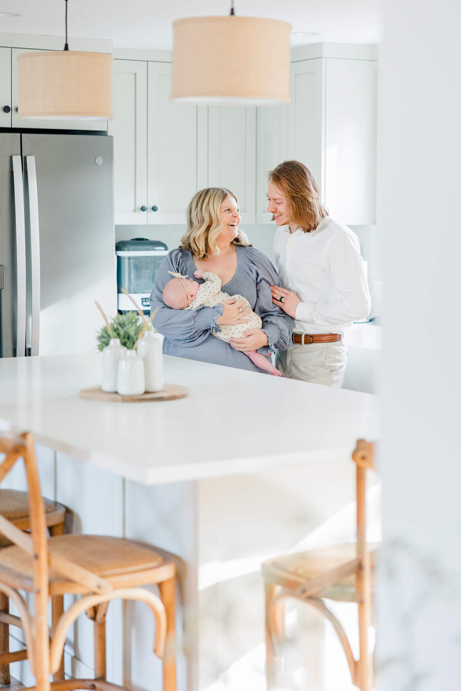 Mom and dad laugh in the kitchen while mom holds their sleeping newborn